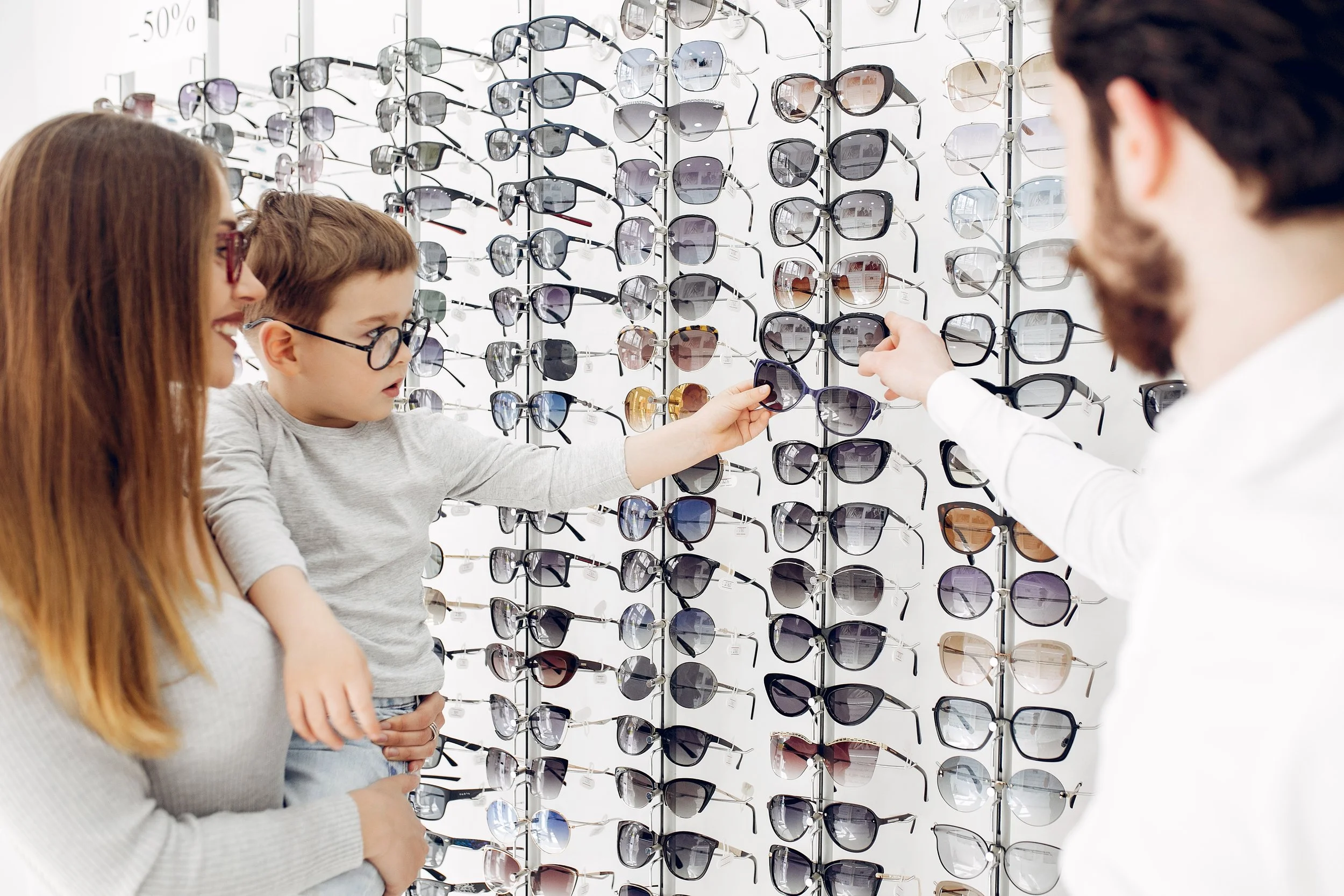 Una mujer, un niño y un hombre observan y eligen gafas de sol en una tienda. La pared detrás está llena de gafas de diferentes estilos y colores.