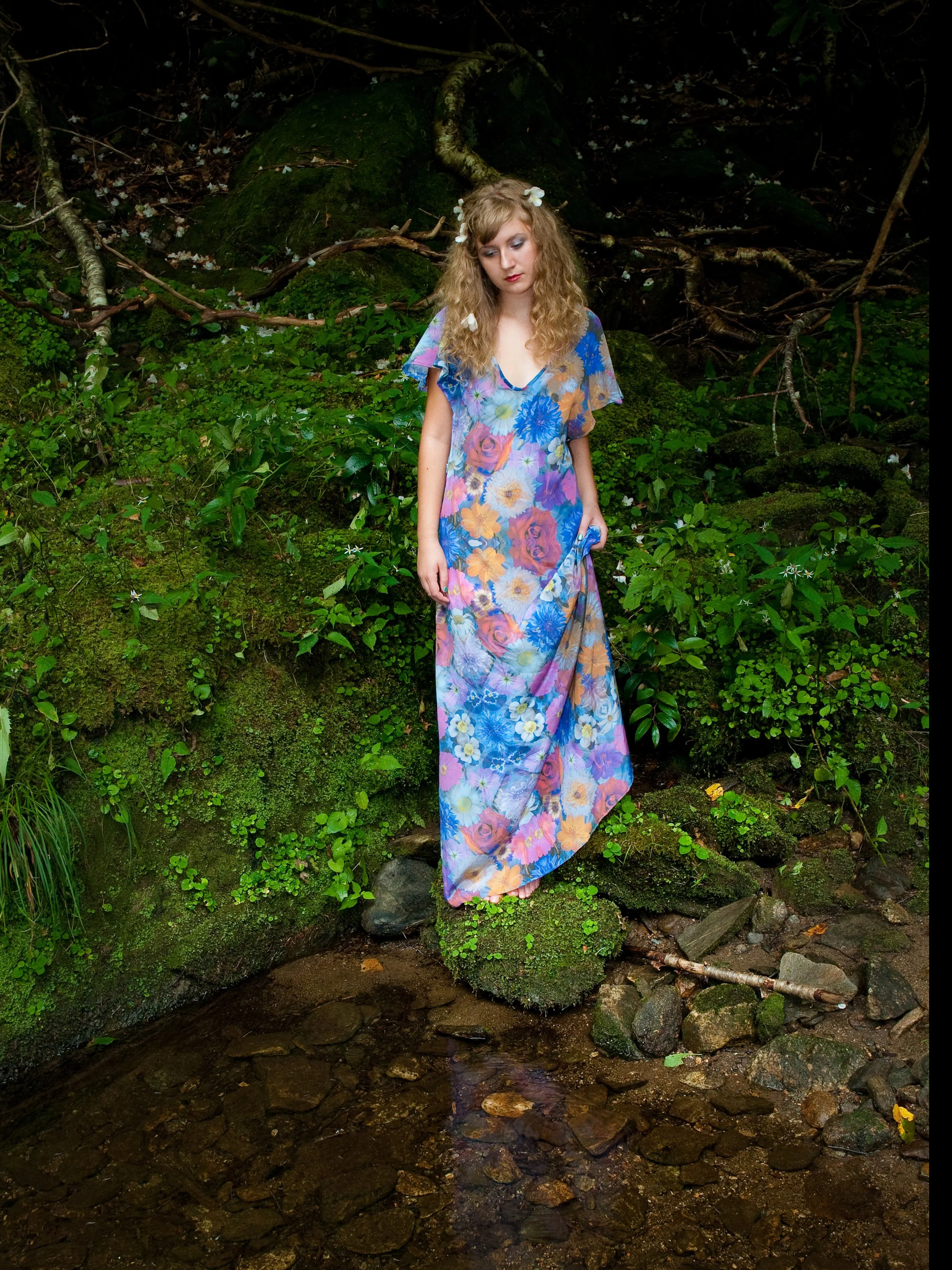 A woman with curly blonde hair, wearing a long, colorful floral dress with a pattern of large flowers, stands in a lush forest near a small stream, surrounded by green foliage and moss-covered rocks. She has a serene expression and a flower in her hair.
