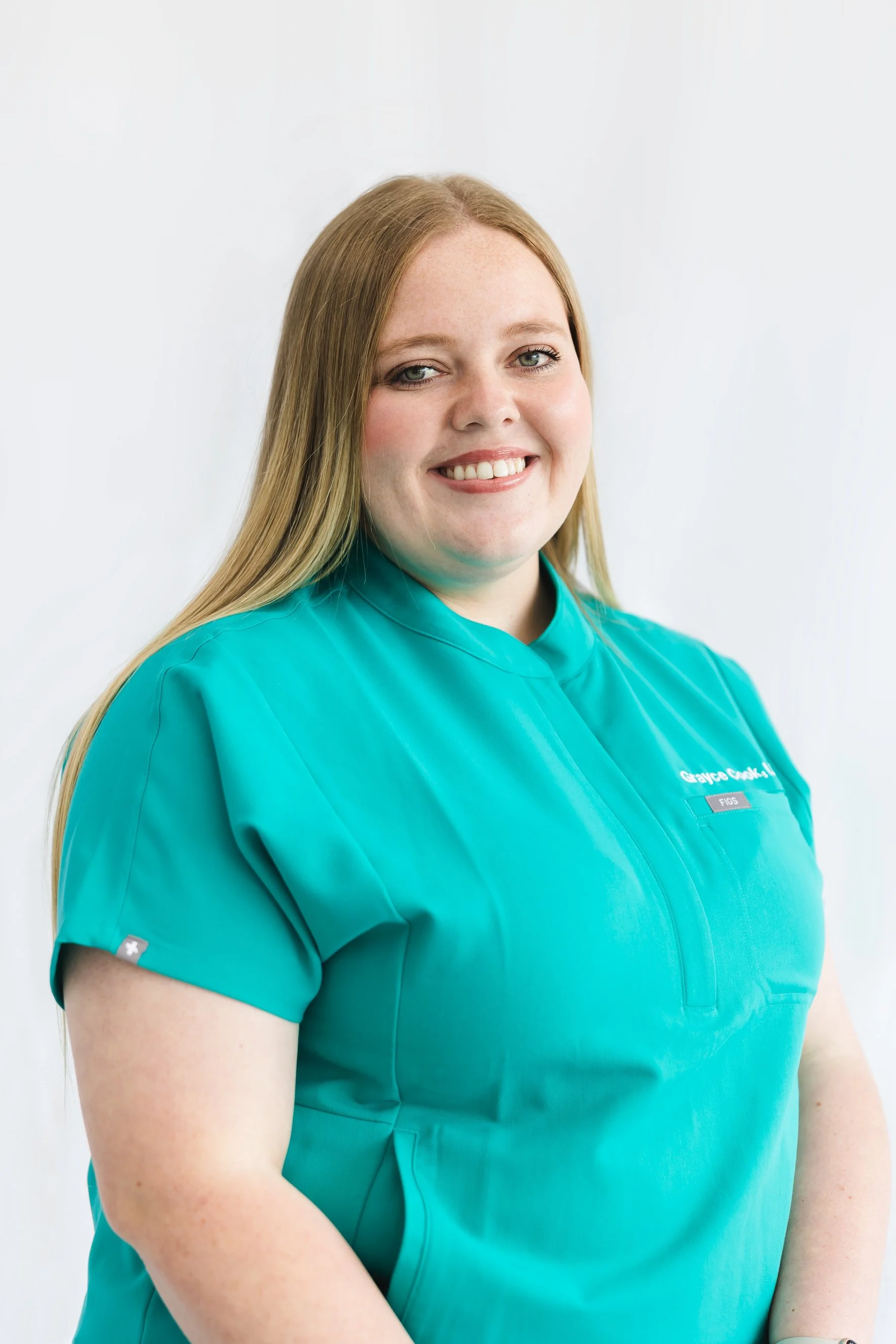 Close-up of a smiling woman with long blonde hair wearing teal medical scrubs standing against a plain white background.