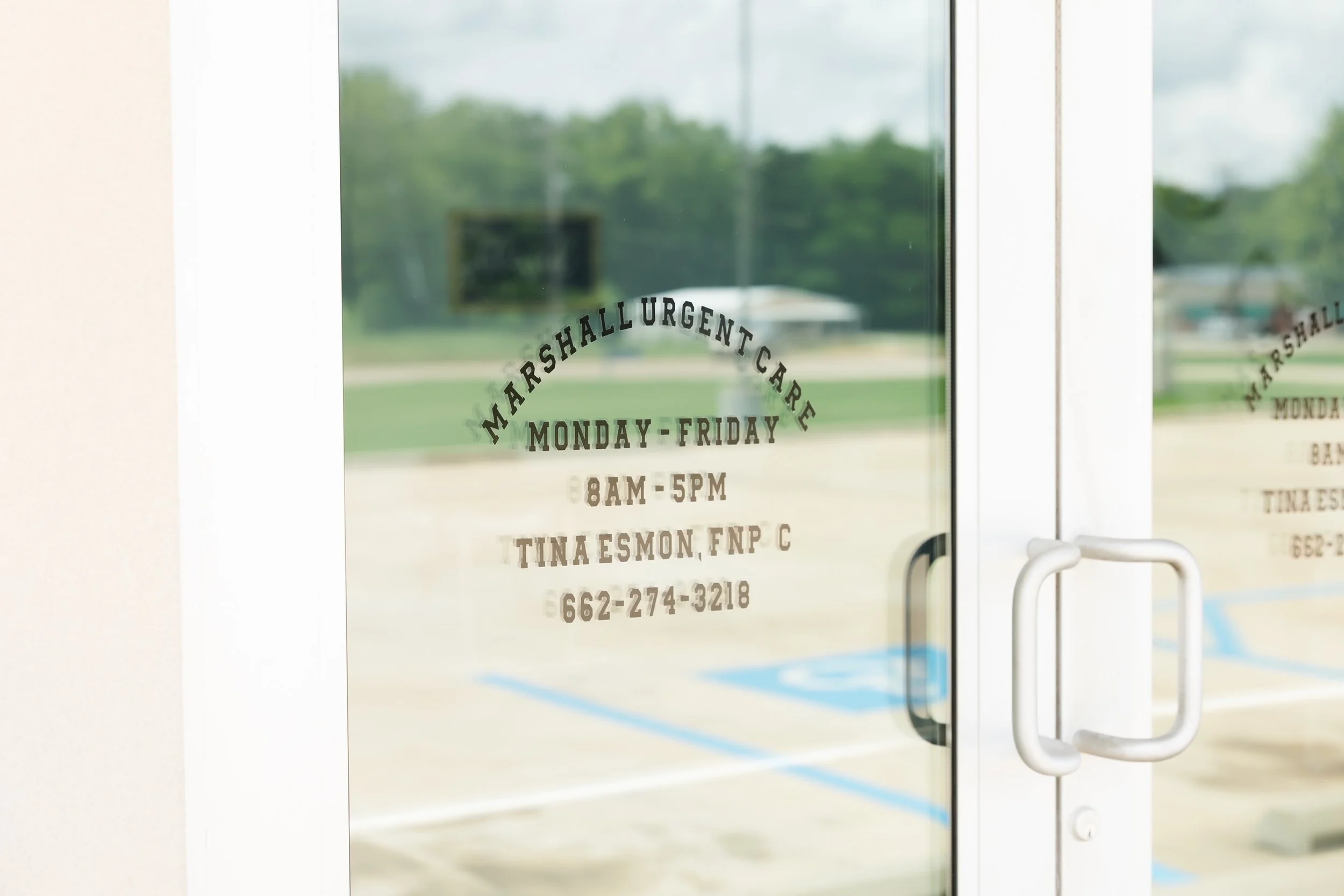 Glass door with a sign displaying the hours of operation for Marshallurgentcare, Monday through Friday from 8AM to 5PM, along with a phone number. Outside view with green trees and a cloudy sky.