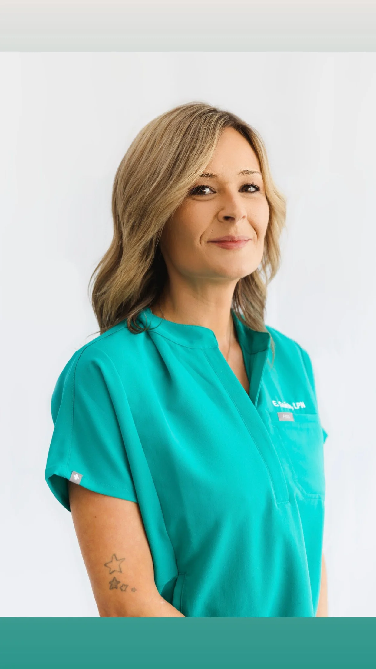 A woman with blonde hair wearing turquoise scrubs, smiling slightly, with a tattoo of stars on her upper arm, standing against a plain white background.