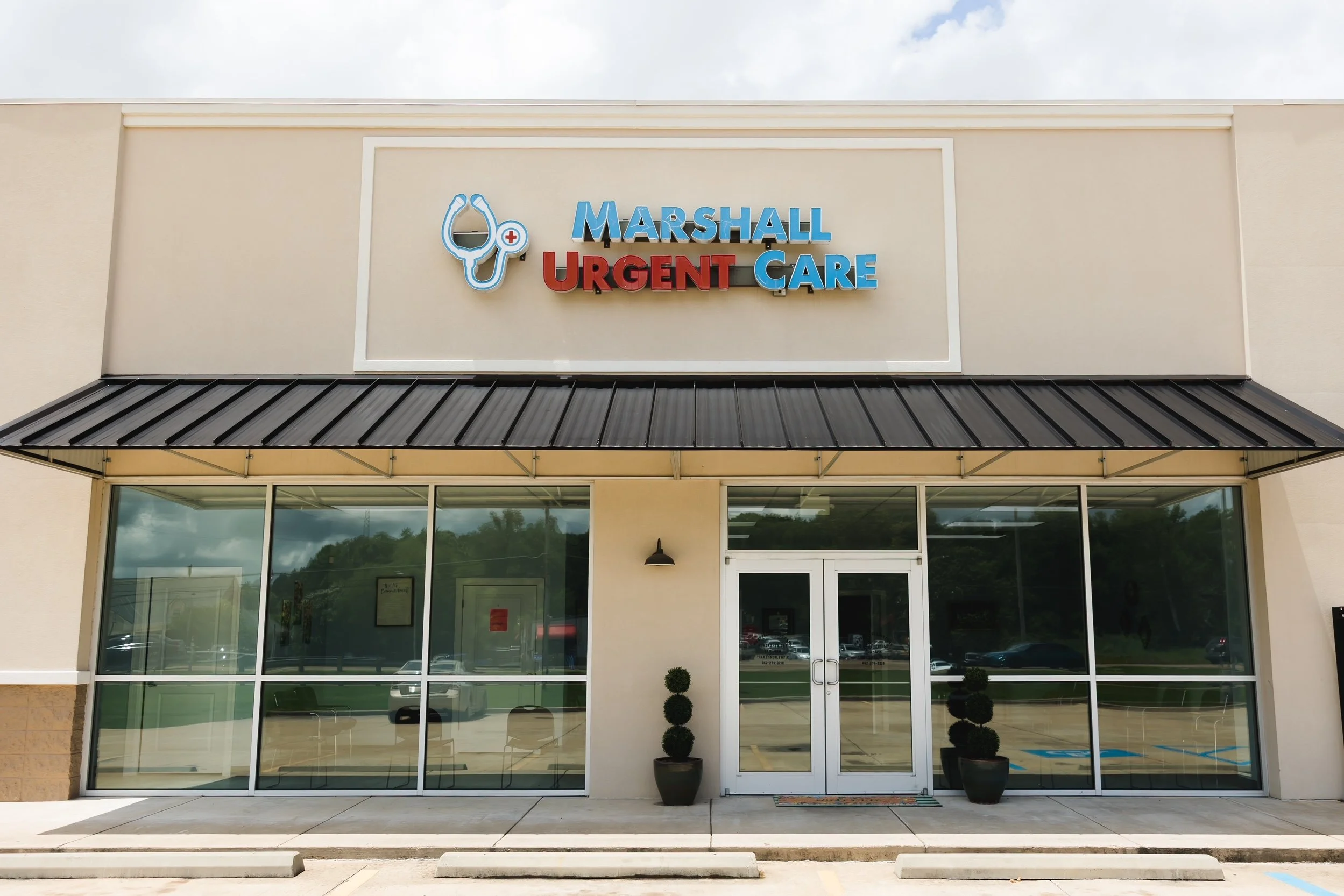 Front view of Marshal Urgent Care building with large glass windows and doors, potted plants, and a sign with a stethoscope graphic and the words 'Marshall Urgent Care' in blue and red letters.