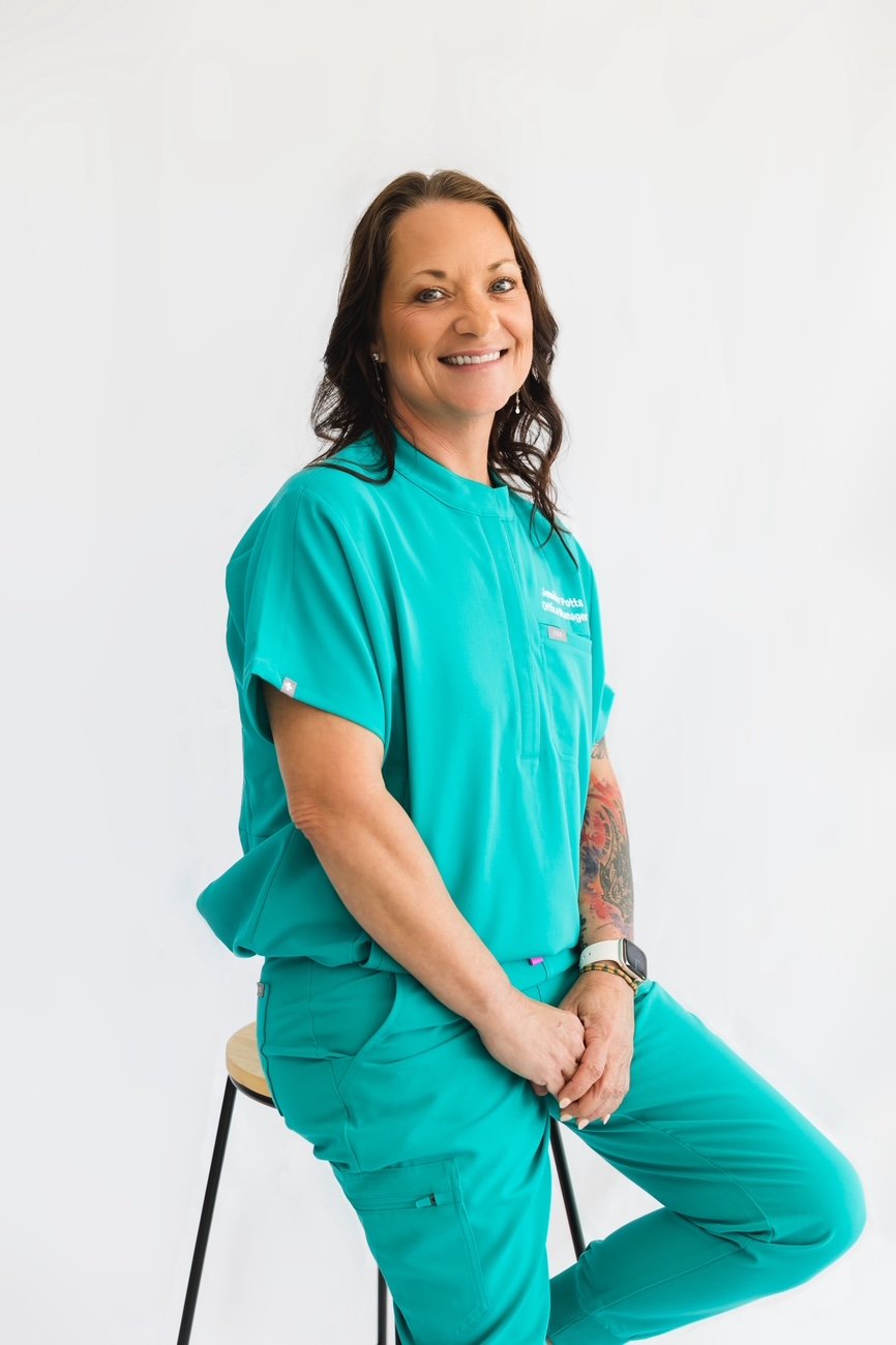 A woman with dark brown hair, wearing teal medical scrubs, sitting on a stool against a white background, smiling at the camera.