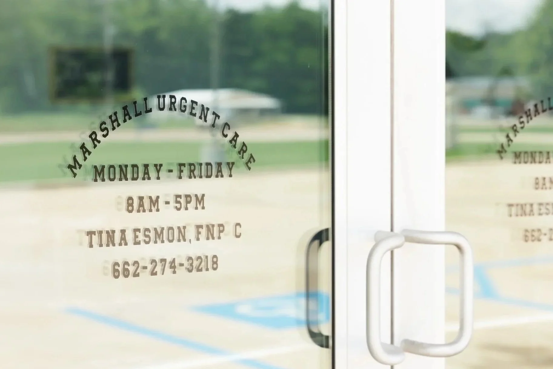 Close-up of a glass door with hours and contact information for Marshallurgent Care, open Monday through Friday from 8 AM to 5 PM, located at Tina Esmon, FNP-C, with a blurred outdoor background.