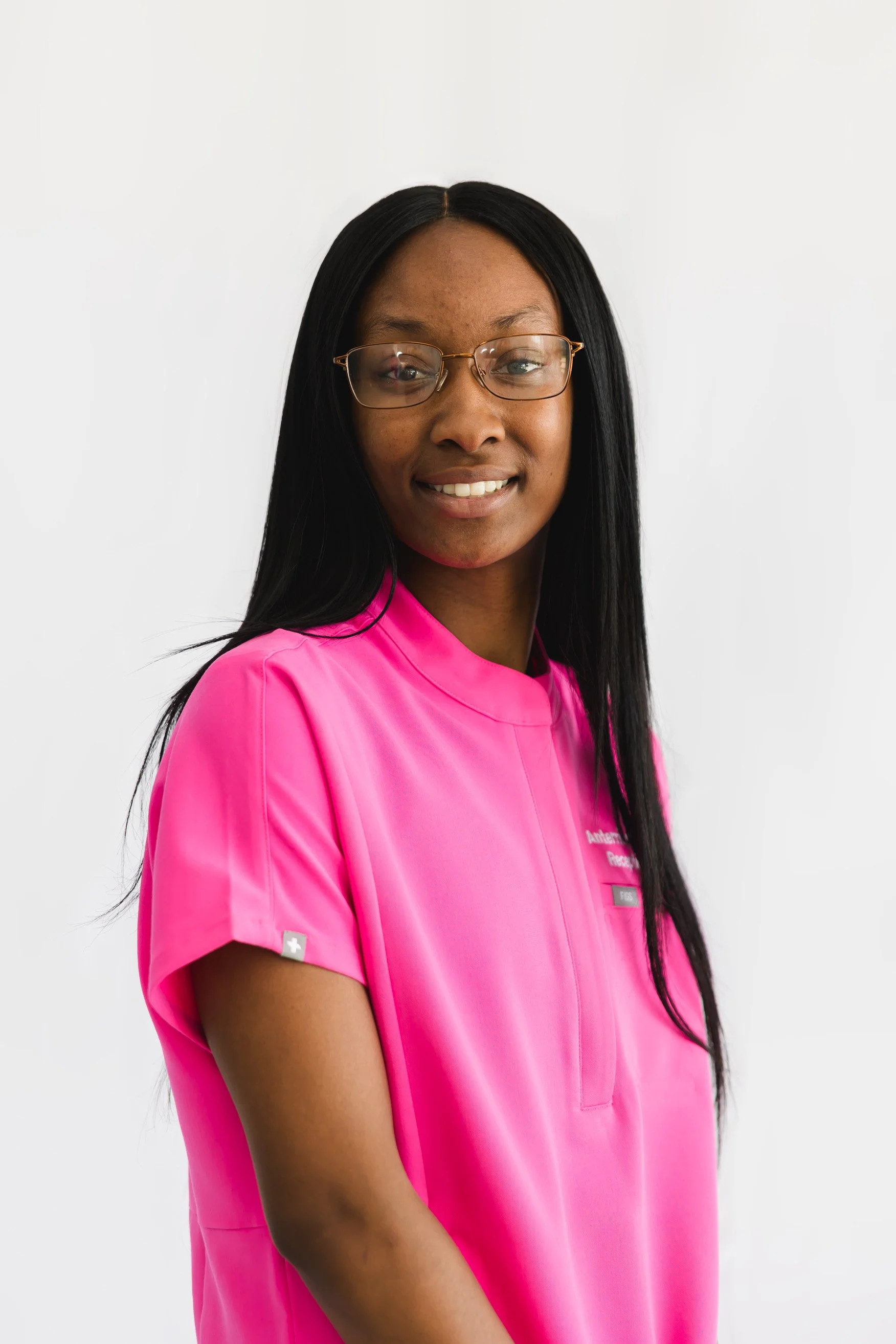 A young African American woman with long black hair, glasses, and a bright pink medical uniform, smiling against a white background.