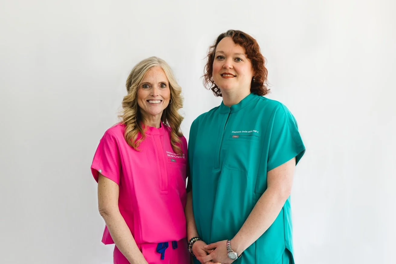 Two women wearing medical scrubs, one in pink and the other in teal, standing next to each other and smiling against a plain white background.