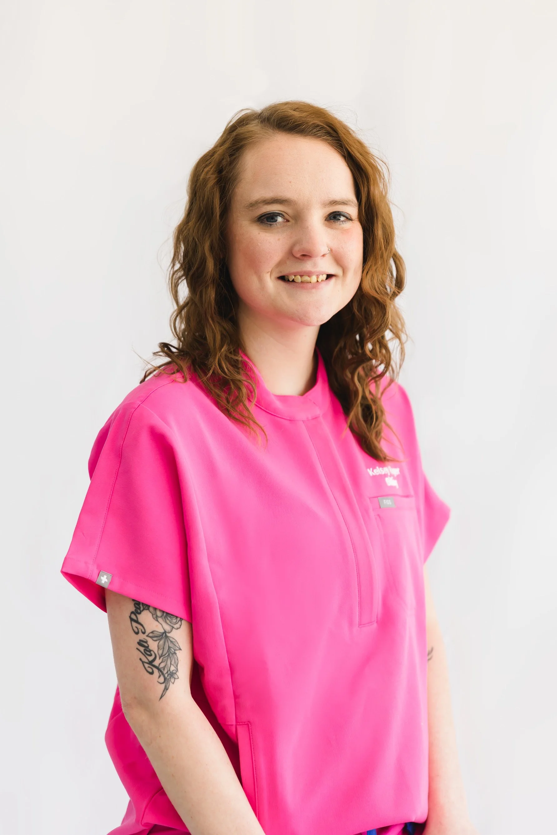 Portrait of a young woman with red curly hair, wearing a bright pink medical scrubs, smiling, against a neutral white background.