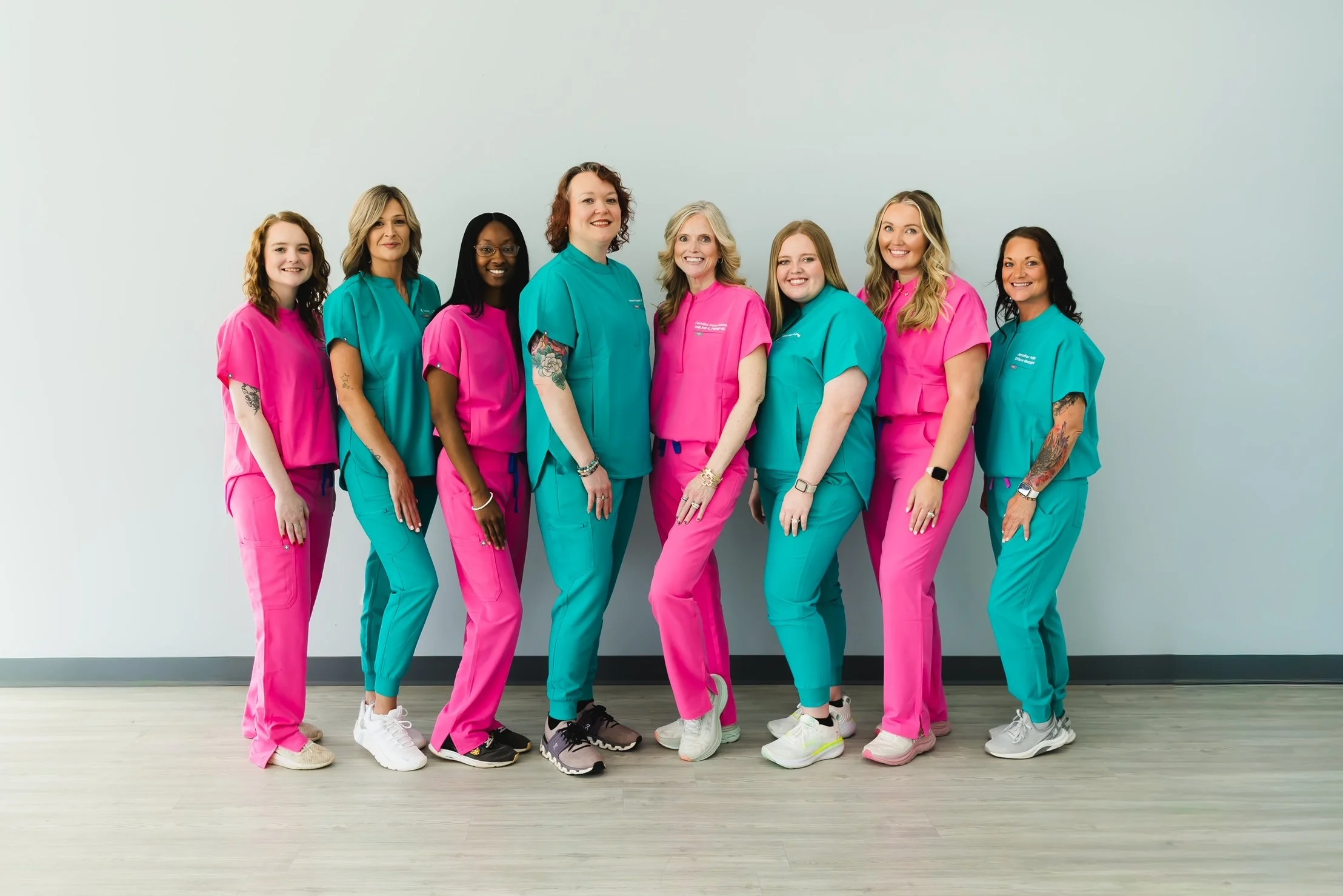 Group of nine women standing in a row against a plain white wall, wearing colorful scrubs and sneakers, smiling at the camera.
