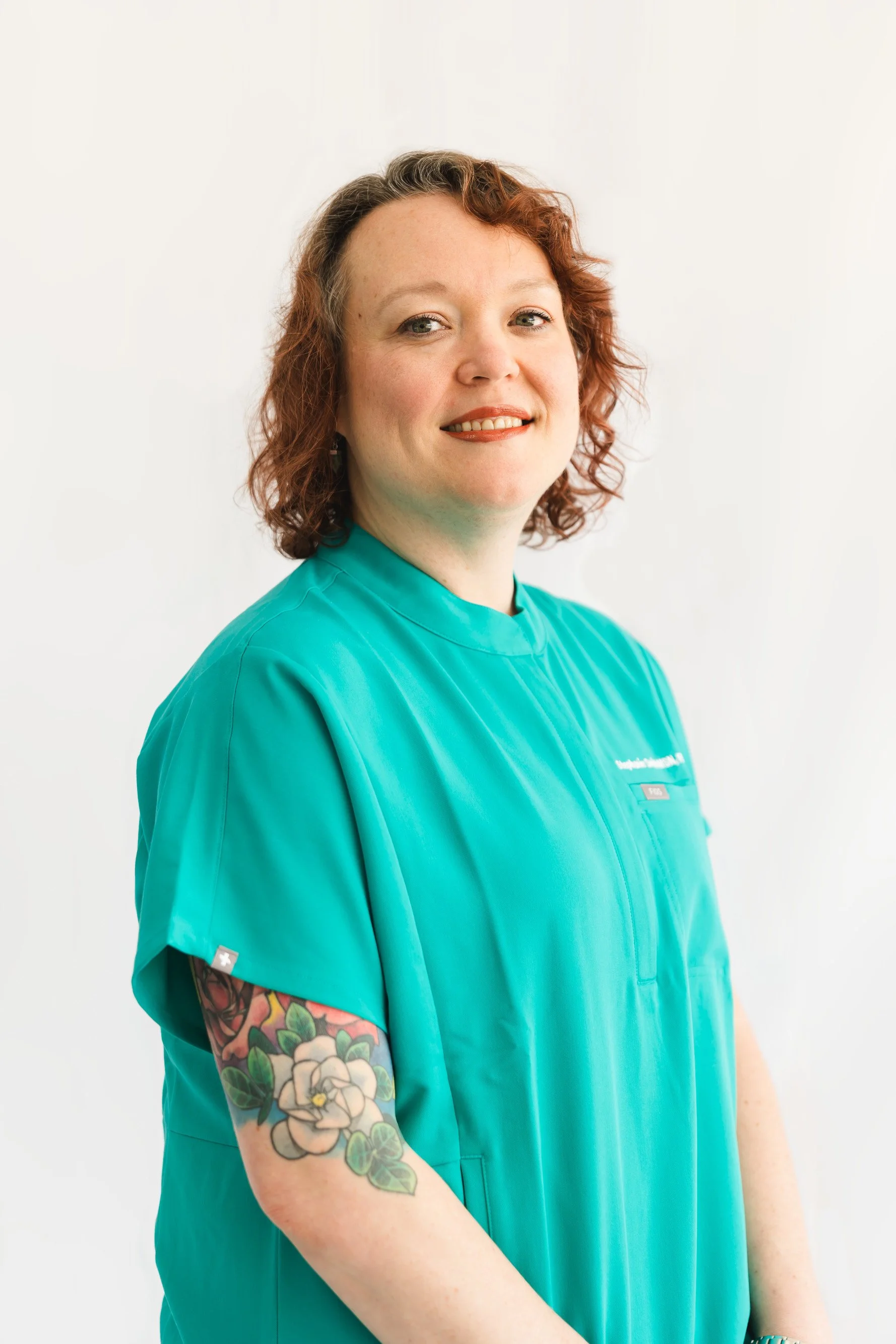 A woman with curly red hair and tattoos on her left arm, wearing teal medical scrubs, smiling and looking at the camera, standing against a plain white background.