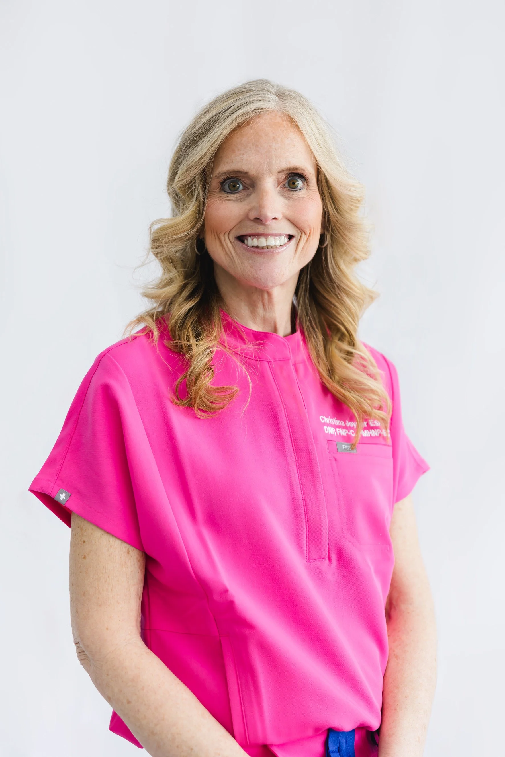 A smiling middle-aged woman with blonde wavy hair wearing a bright pink medical scrub top, standing against a plain white background.