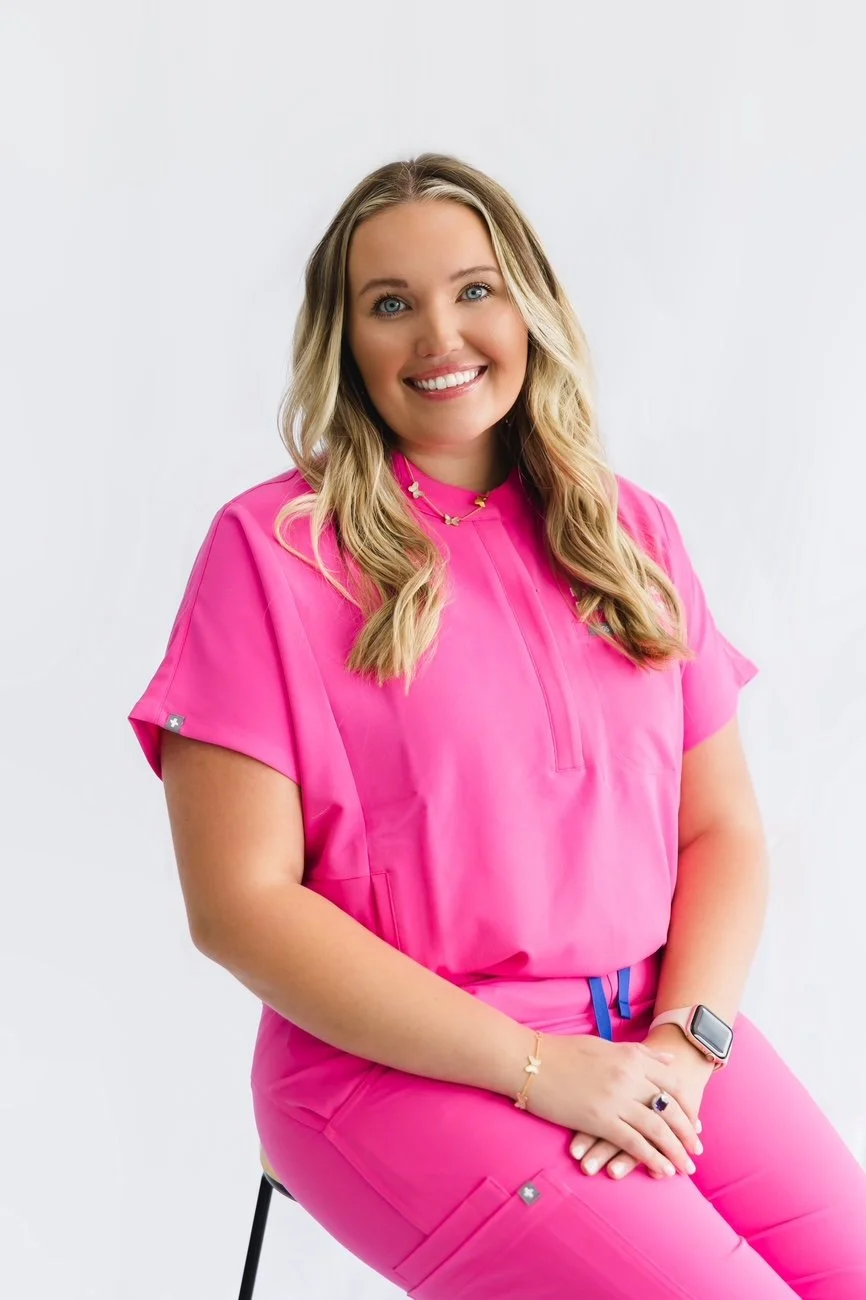 A woman with long blond hair, blue eyes, and fair skin, smiling, wearing a pink medical uniform and sitting on a stool against a plain white background.