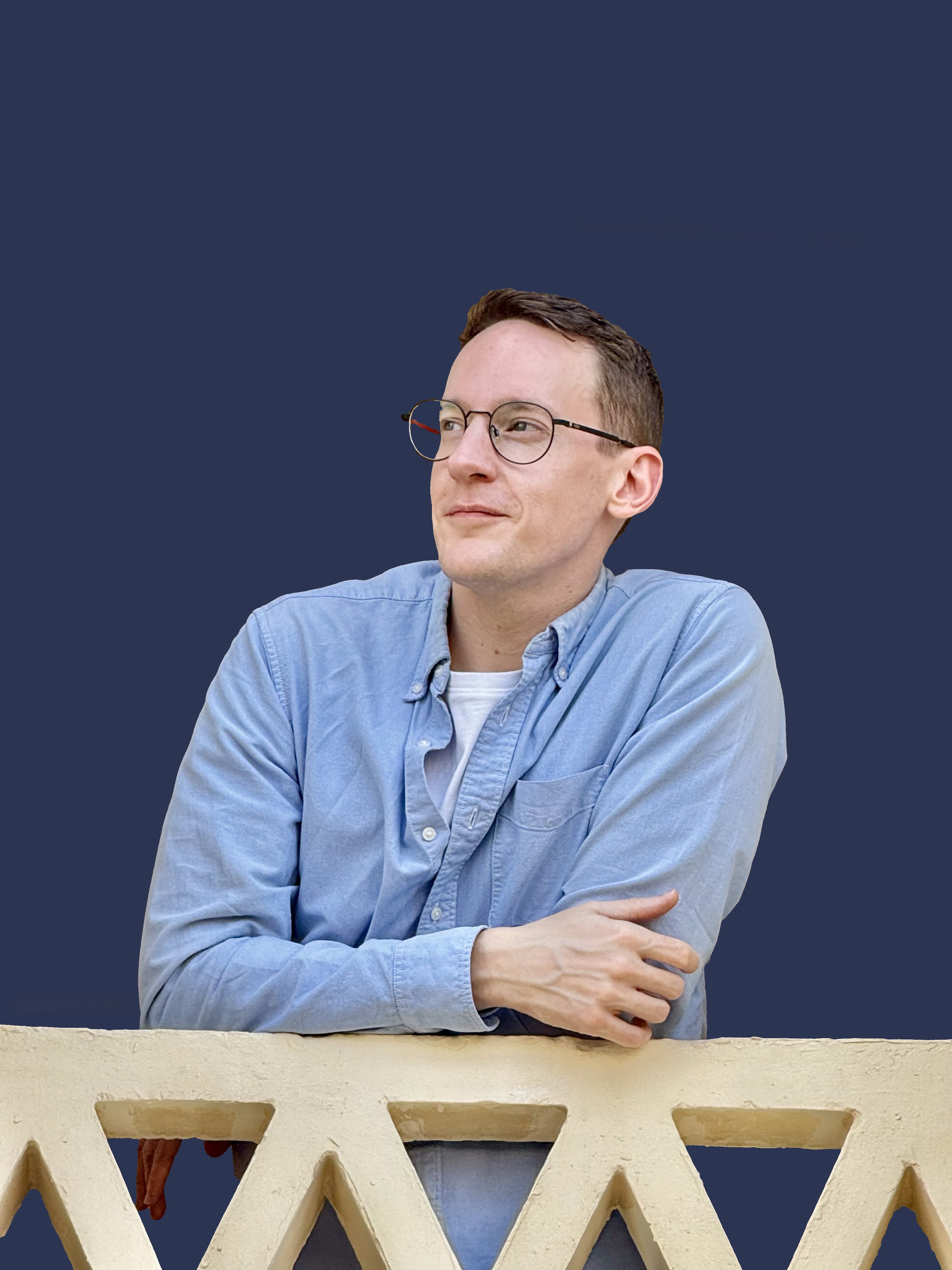 A young man with short brown hair, glasses, and a light blue shirt sitting behind a decorative white railing against a navy blue background.