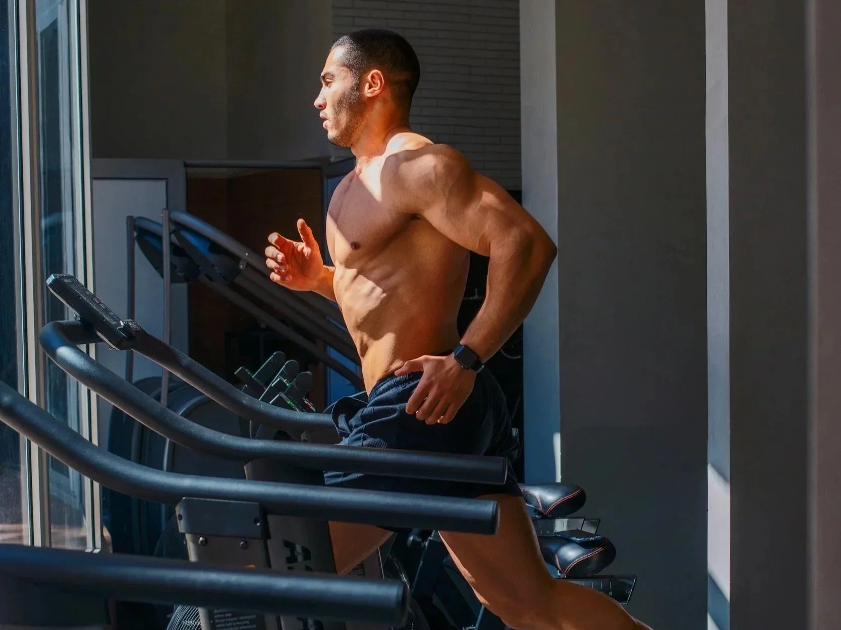 A shirtless man running on a treadmill in a gym or fitness center.