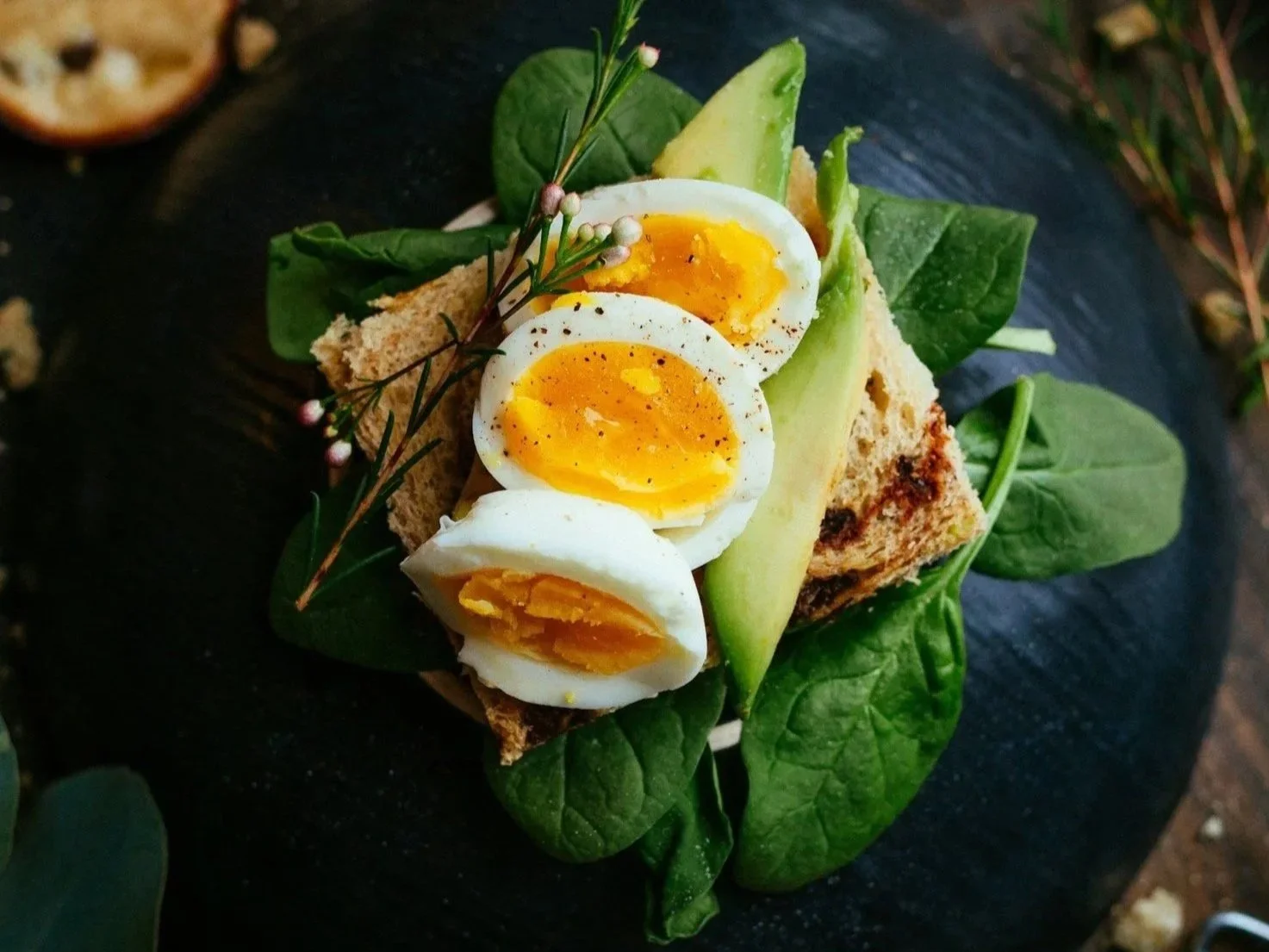 A sandwich with three halved soft-boiled eggs, leafy greens, a slice of avocado, and herbs on a dark surface.