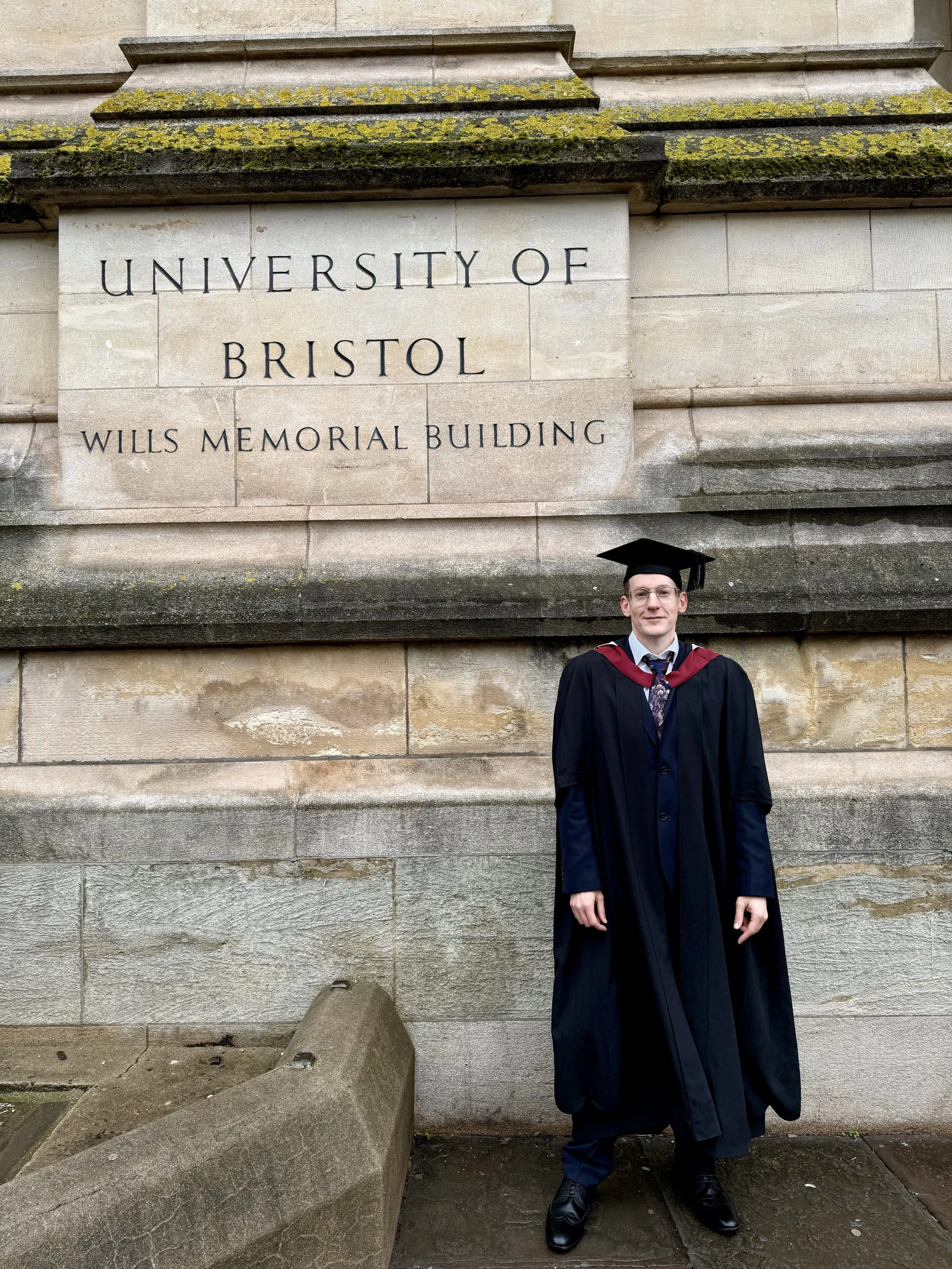 A young man in a graduation cap and gown standing in front of a stone wall that reads 'University of Bristol Wills Memorial Building'. Various moss and lichen grow on the wall, and there is a large stone sculpture at his feet.