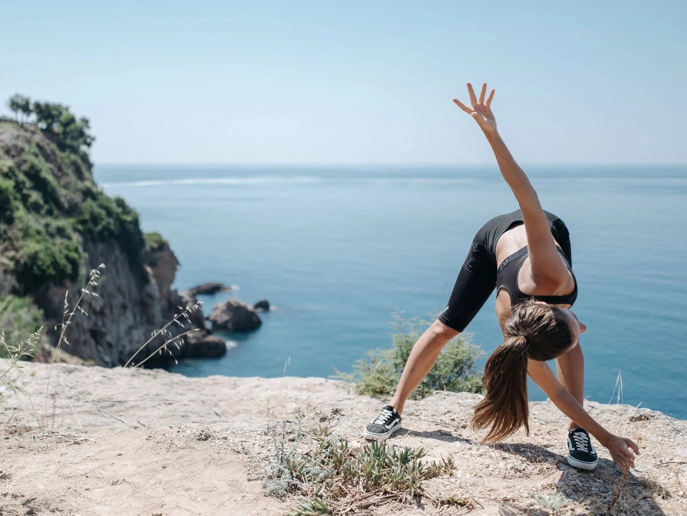 A woman practicing yoga on a rocky beach with a cliff and ocean in the background, performing a backbend pose with her arms extended upward.