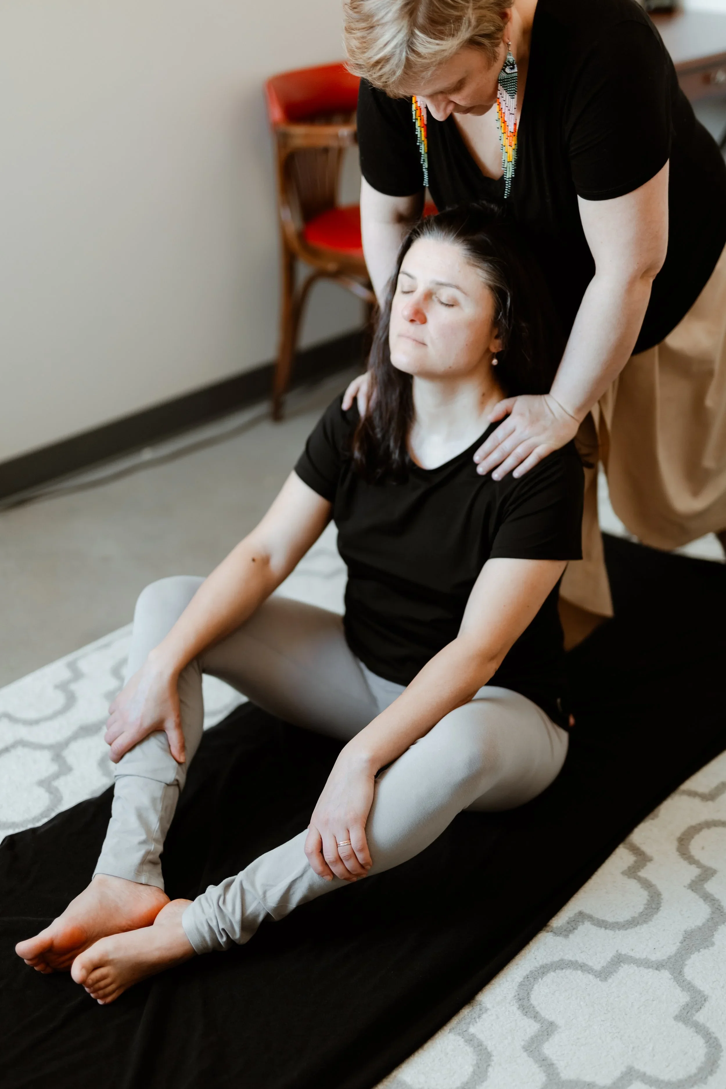 Une femme semble recevoir un massage ou une thérapie, avec une autre femme positionnée derrière elle, dans une pièce intérieure.