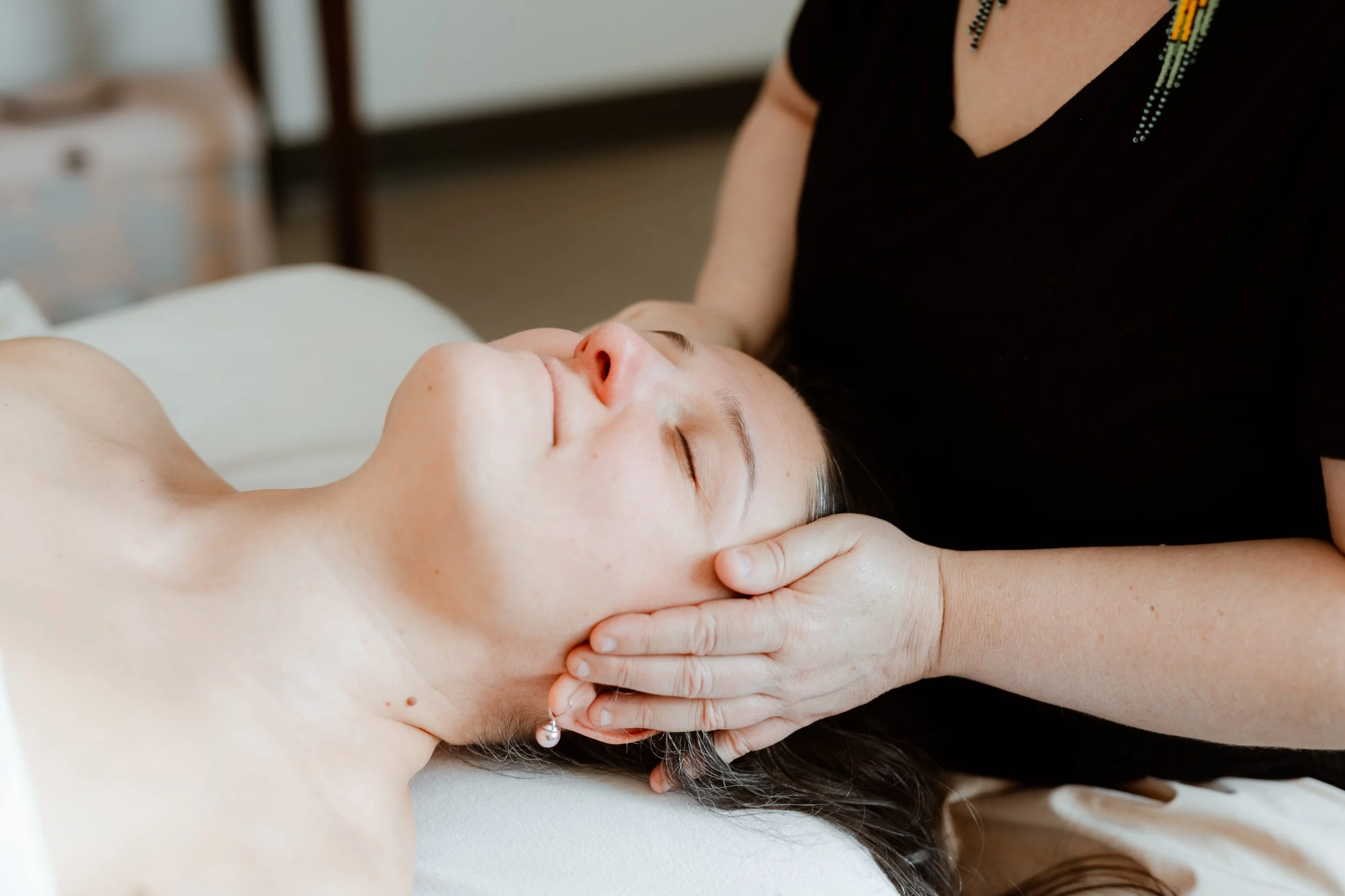 Une femme reçoit un massage ou une séance de soins du visage, allongée sur une table, les yeux fermés, une autre personne lui tient la tête et la nuque pendant qu'elle repose.