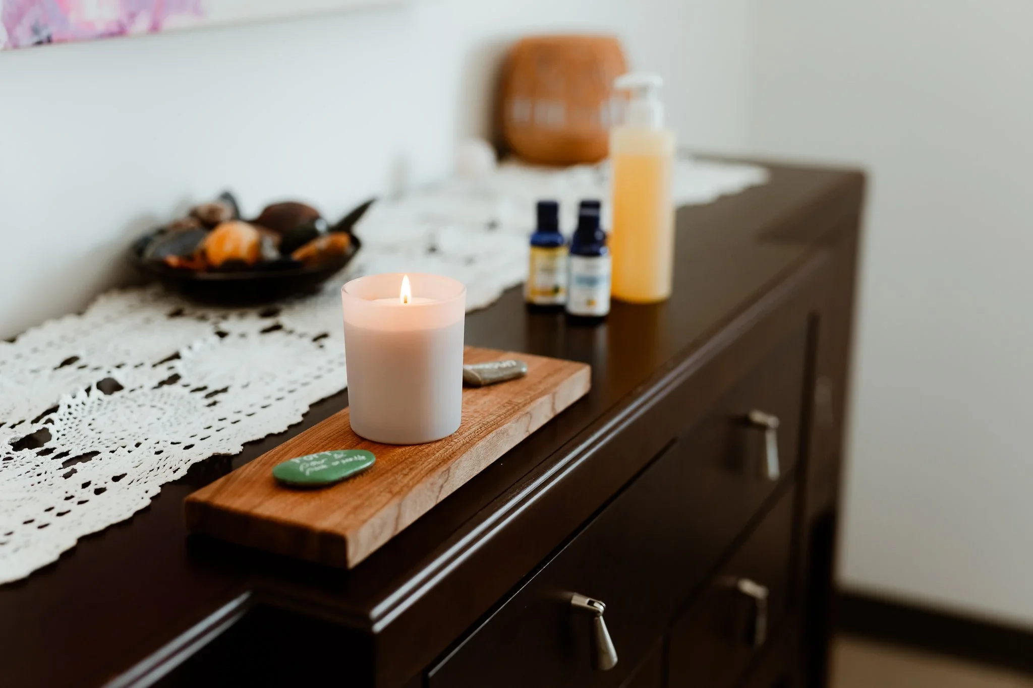 Bougie blanche allumée sur une planche en bois posée sur une commode en bois foncé, avec des huiles essentielles et un pot en terre cuite sur une nappe en dentelle blanche, dans une pièce au mur blanc.
