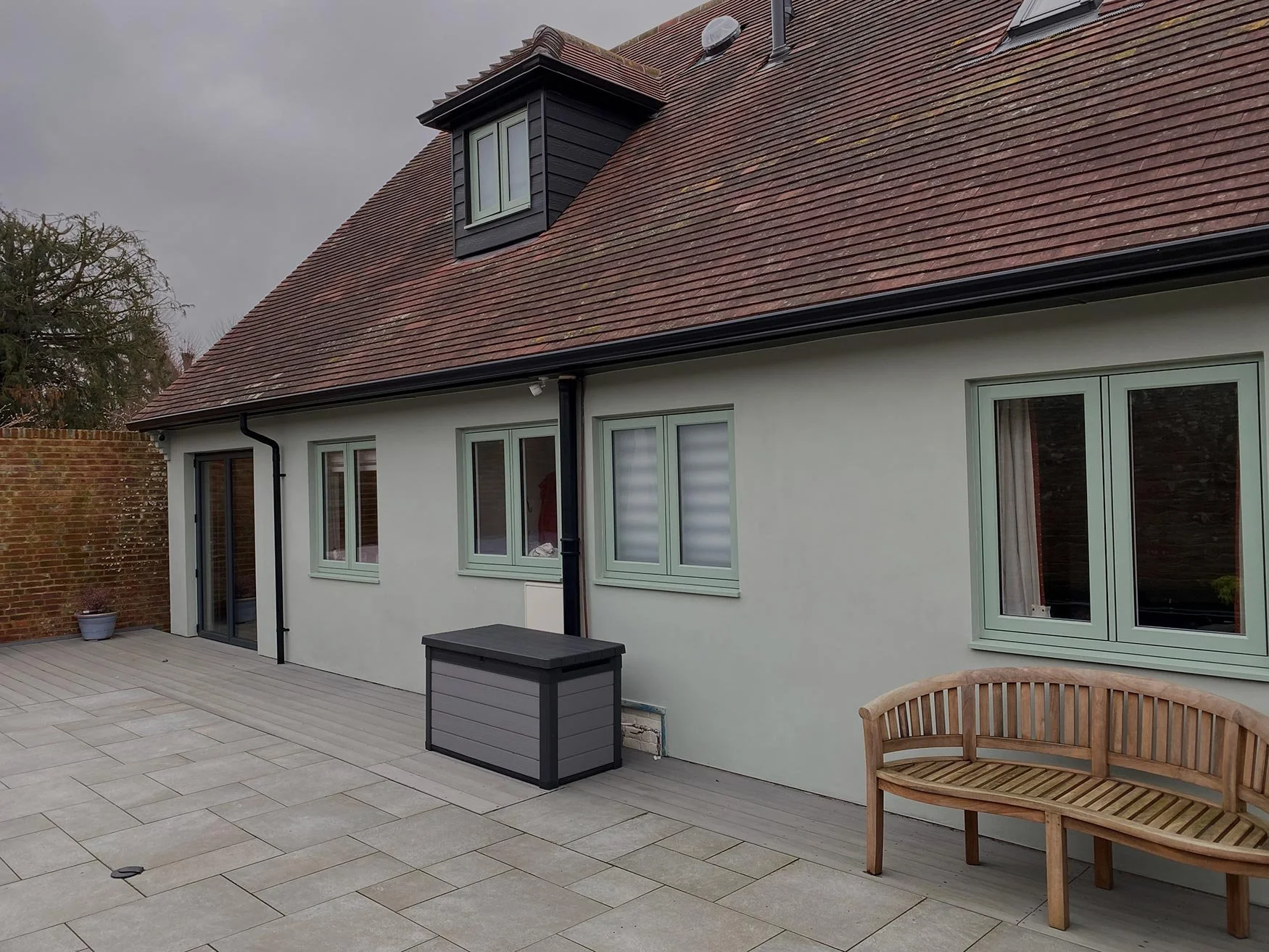 Backyard patio with large light-colored stone tiles, a gray storage box, a wooden bench, and a white house with multiple windows and a red tiled roof. The backyard is enclosed by a brick wall, and there is a potted plant near the wall. Overcast sky.