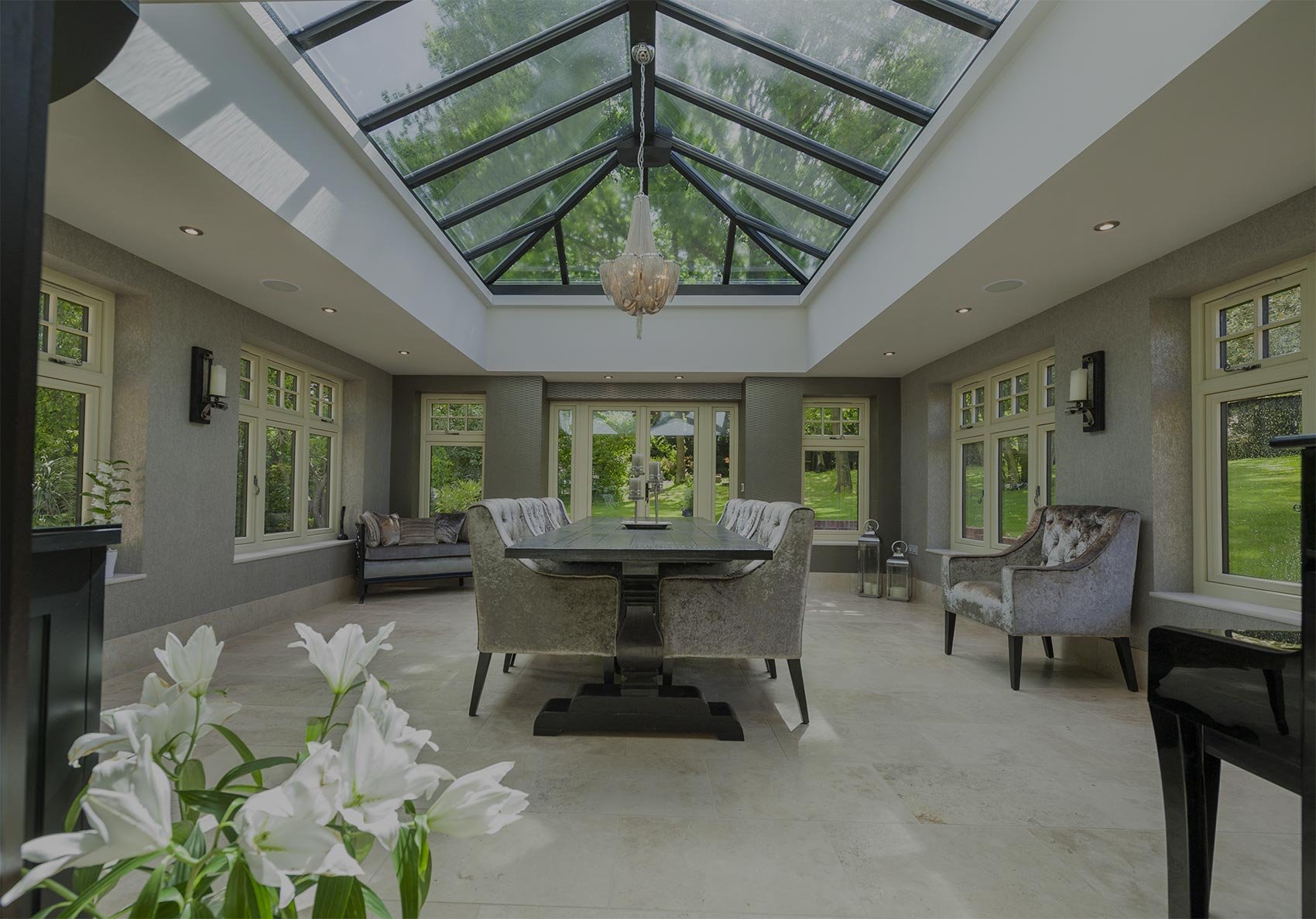 A bright sunroom with a large rectangular dining table surrounded by velvet chairs, illuminated by a glass ceiling chandelier and natural light from multiple windows, overlooking a lush green yard.