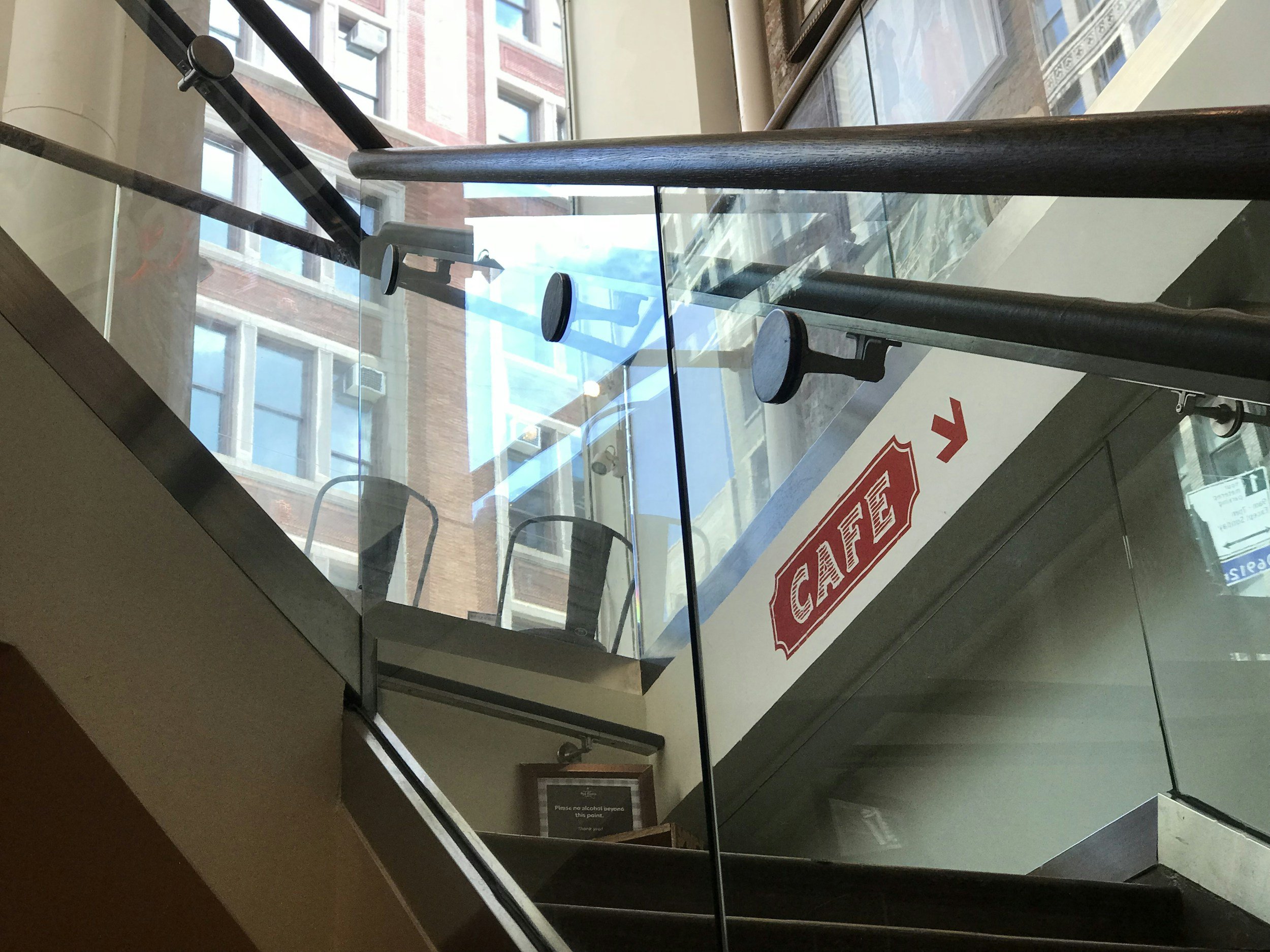 Interior view of a staircase with glass railing, overlooking a city street with brick buildings and windows.