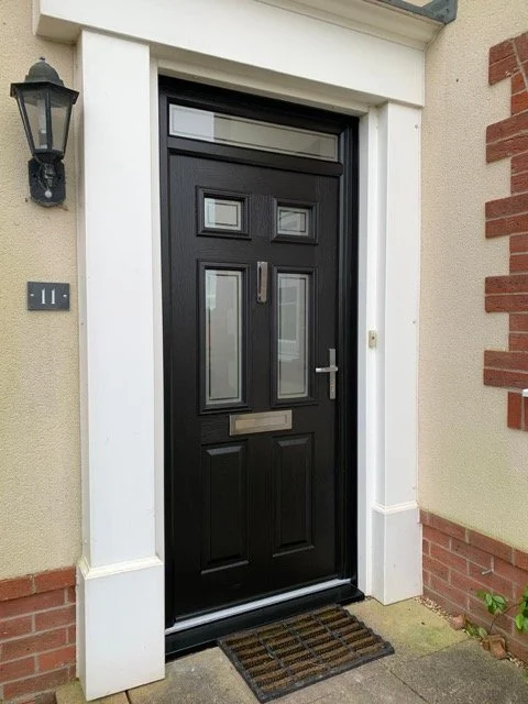 Black front door with a mail slot, silver handle, and glass panels, surrounded by white trim, with a lamp fixture on the exterior wall and a welcome mat in front.