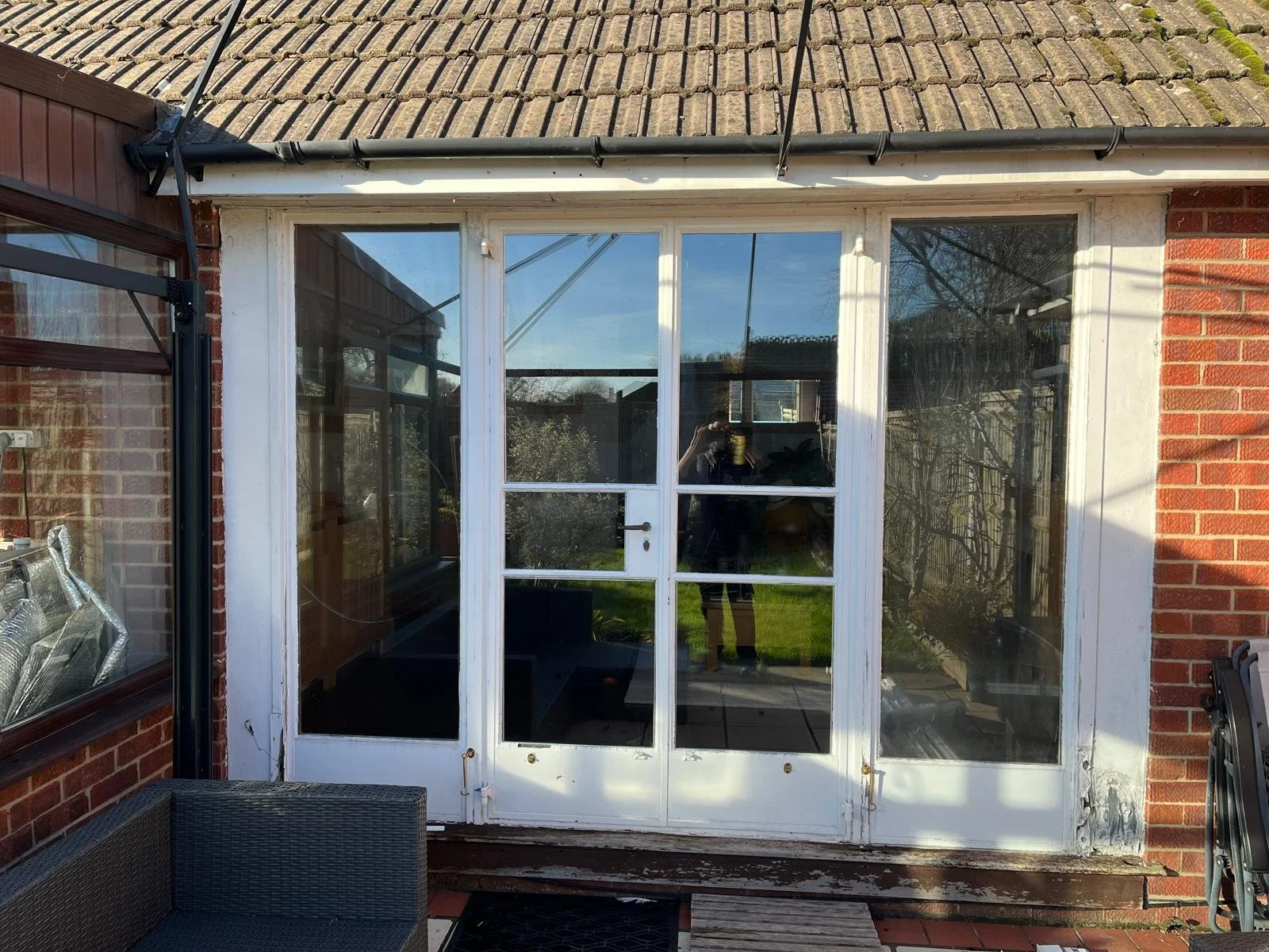 Outdoor view of a house's back door with large glass panes, surrounded by red brick walls and a tiled roof. Reflection of a person taking the photo is visible in the glass.