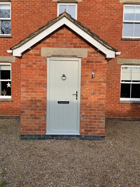 A small brick structure with a light gray door, a door knocker, a mail slot, and a latch, attached to a larger brick building with three windows visible.