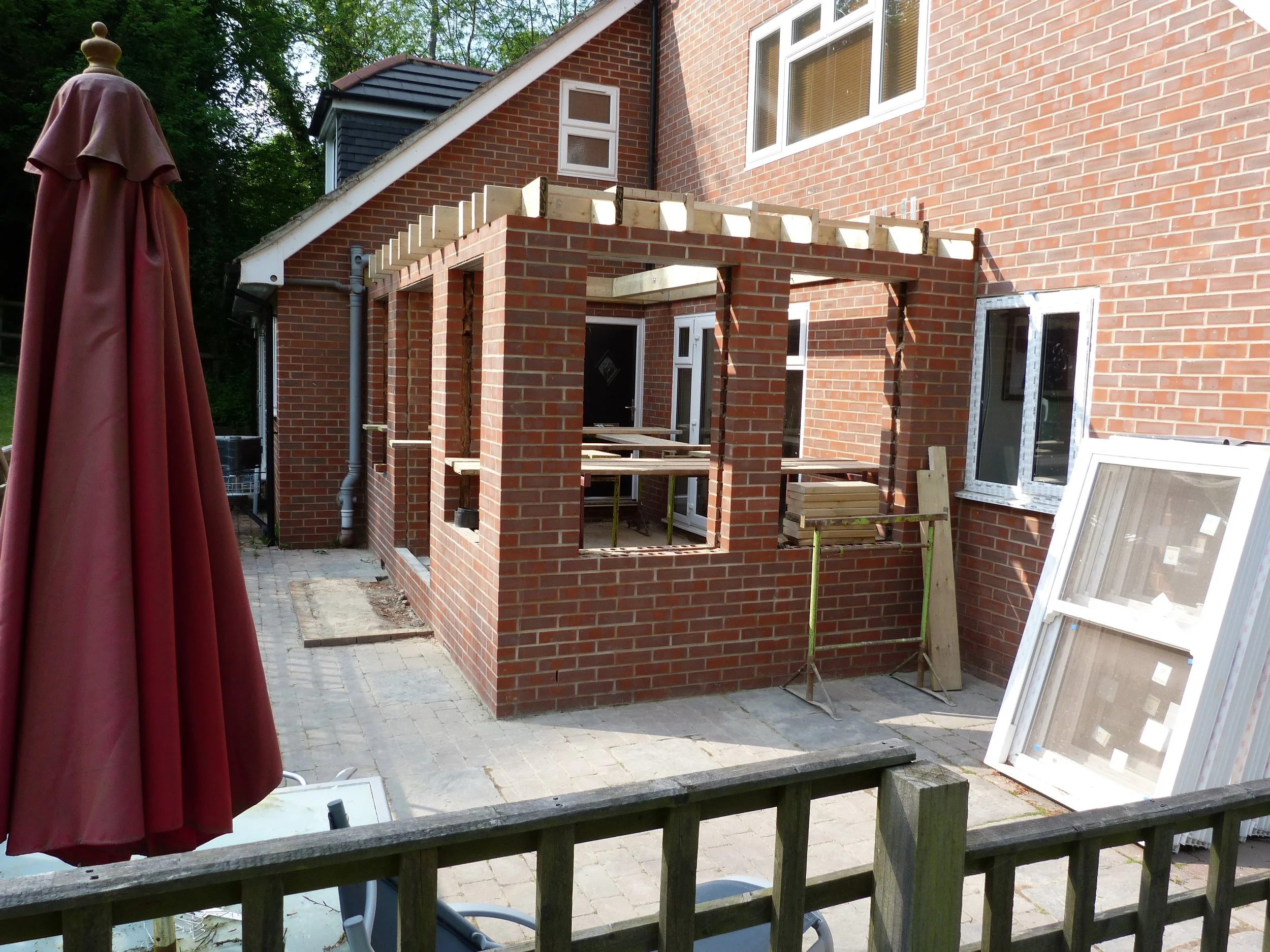 An outdoor scene of a brick house under construction with a partially built brick frame for a new addition, a closed patio umbrella on the left, and windows on the house. The area is paved with stone bricks.