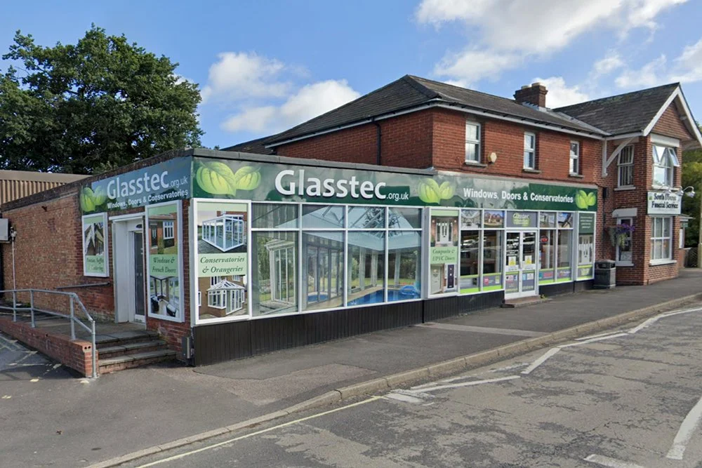 Exterior of a glass and door shop with a green sign reading 'Glasstec' and advertisements for windows, doors, conservatories, and orangery products.