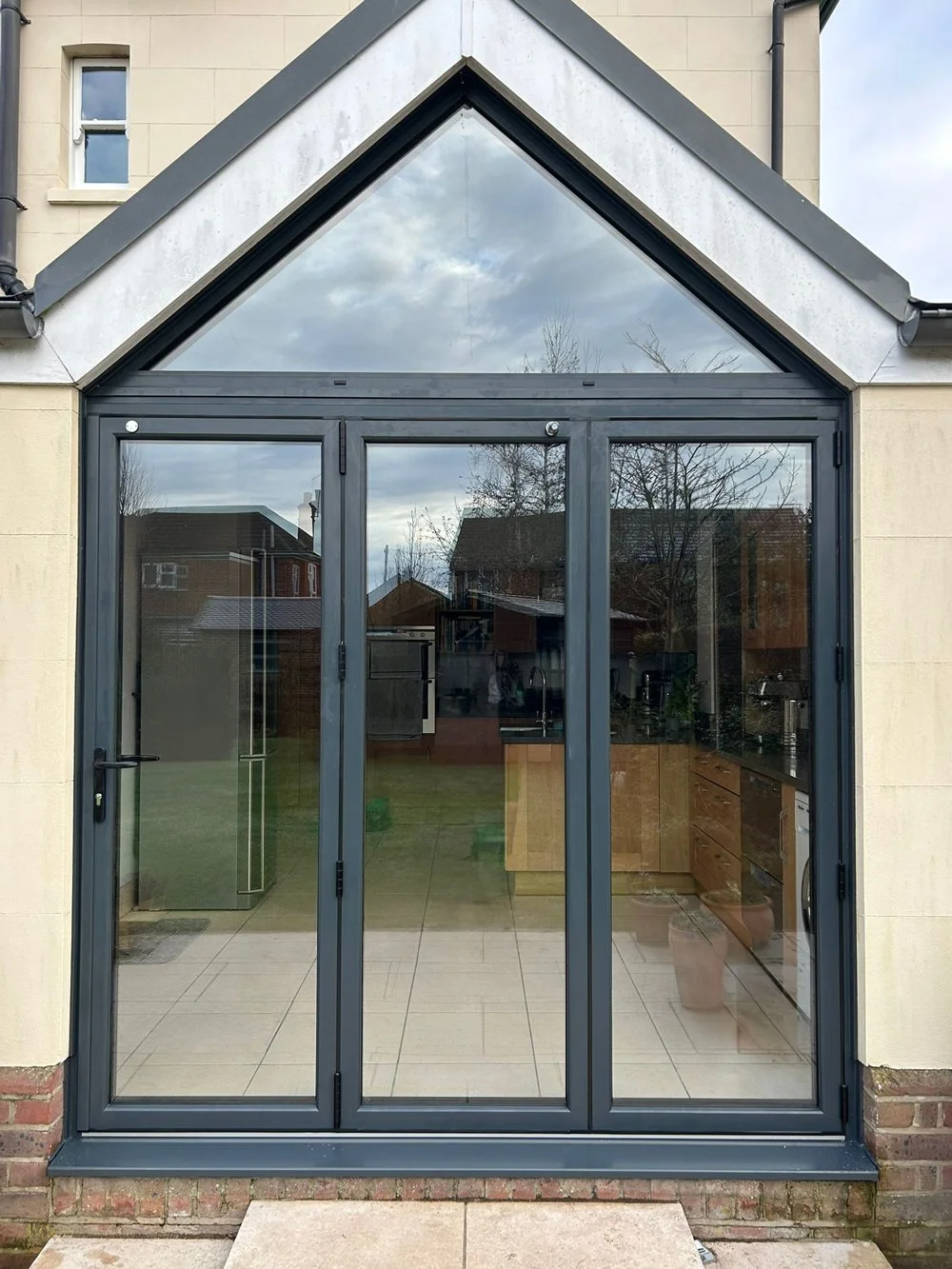 A large glass door with multiple panels, leading into a kitchen with wooden cabinets and a tiled floor, reflected in the glass are houses, trees, and a cloudy sky.