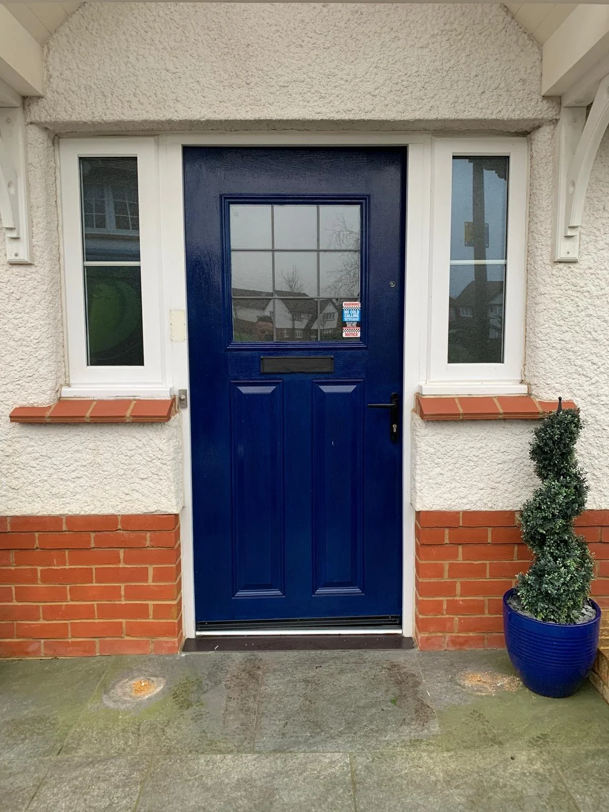 A blue front door with a window, surrounded by two small windows, with white trim, on a house with beige textured walls and a brick bottom border. There are brick window sills, and a potted bush with a blue pot on the right side.