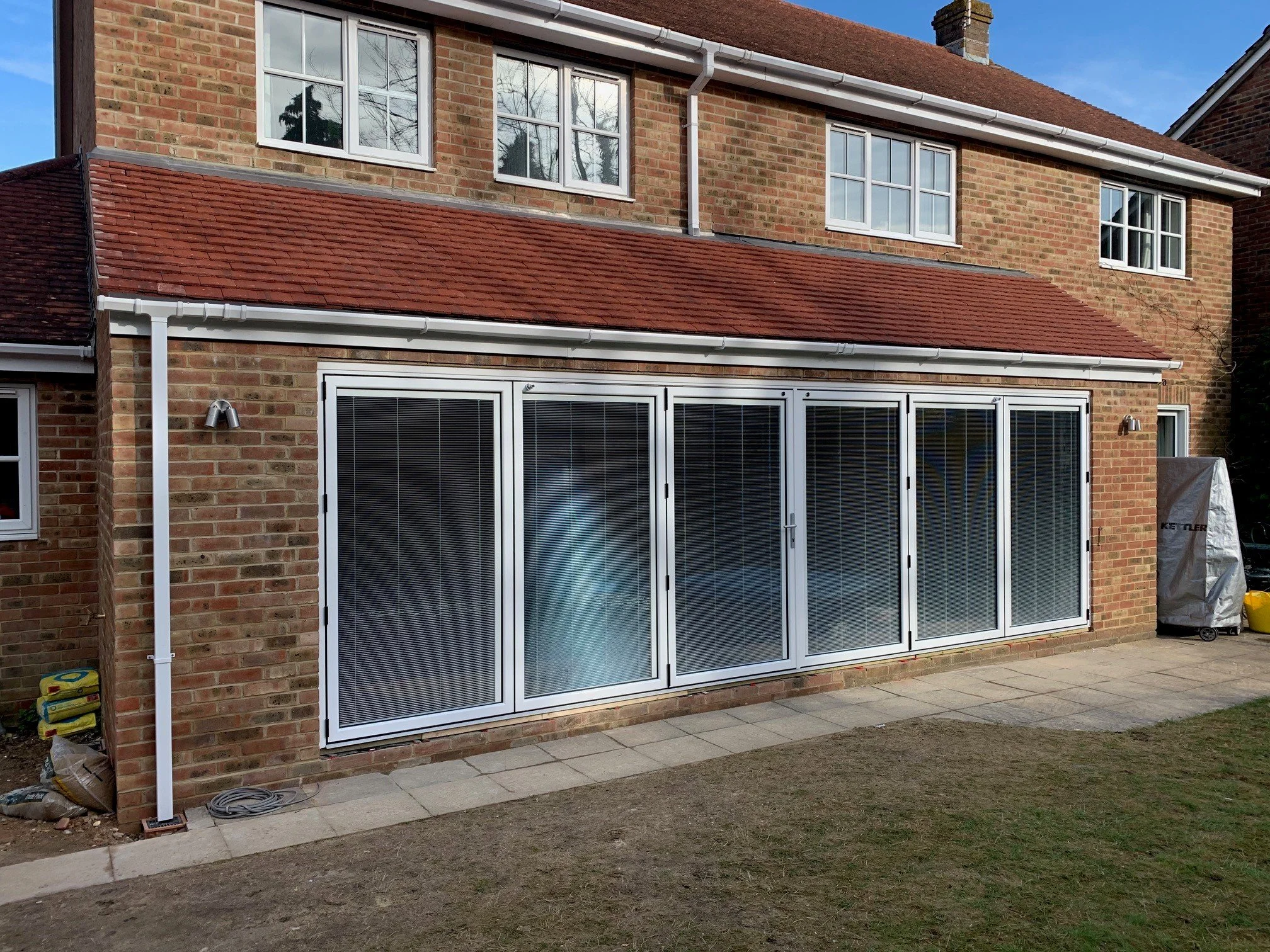 Back view of a brick house with large glass sliding doors and elevated windows, red tiled roof, and outdoor patio area with paving stones, some garden tools and materials on the ground.