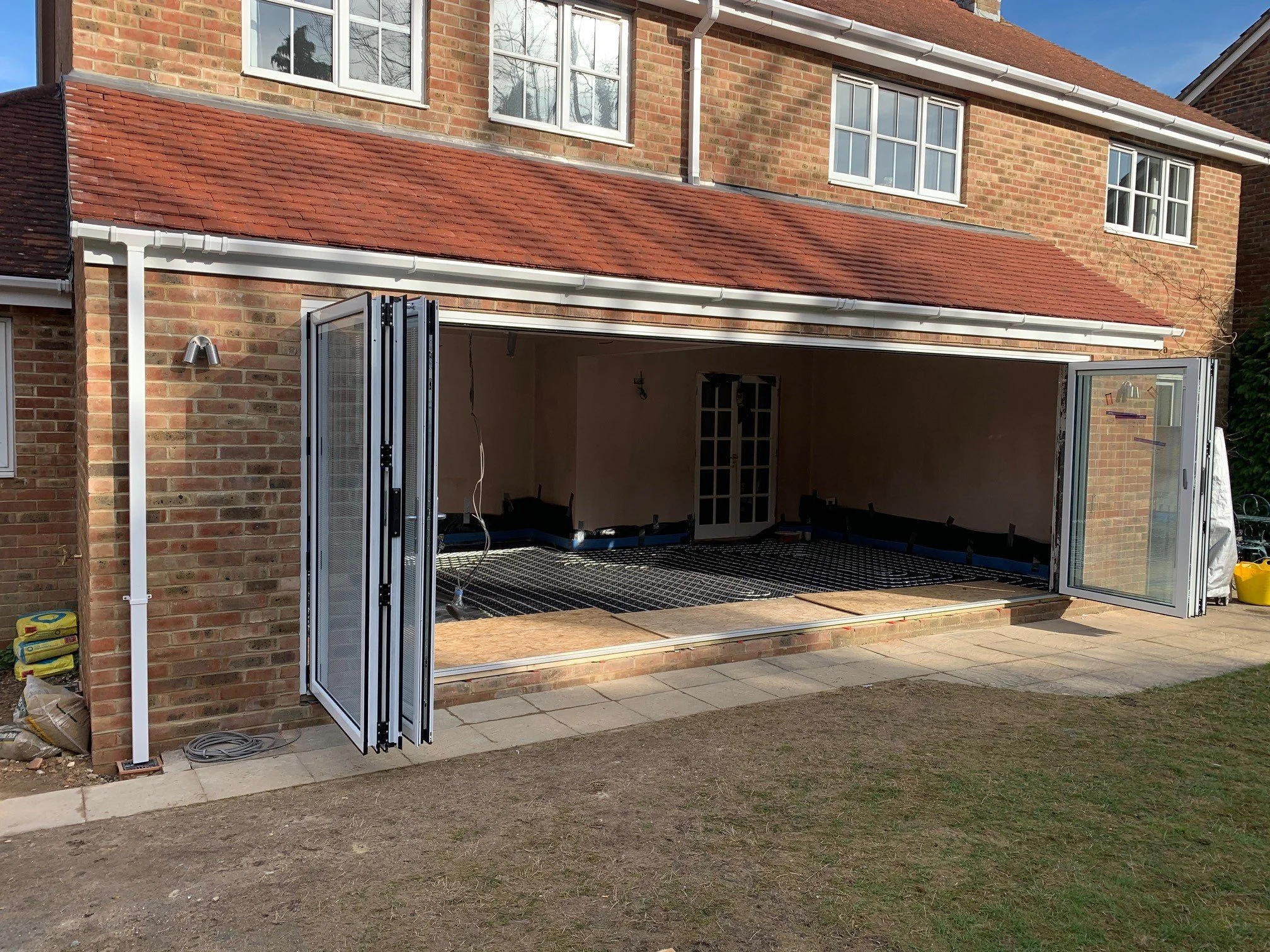 A brick house with large windows and an open garage with folding glass doors. The garage is under construction, with construction materials and tools visible inside. There is a paved patio in front and a patch of grass and soil nearby.