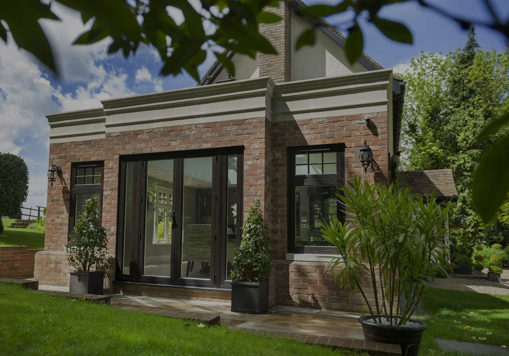 Back view of a brick house with large glass doors, potted plants, trees, and a green lawn under a partly cloudy sky.