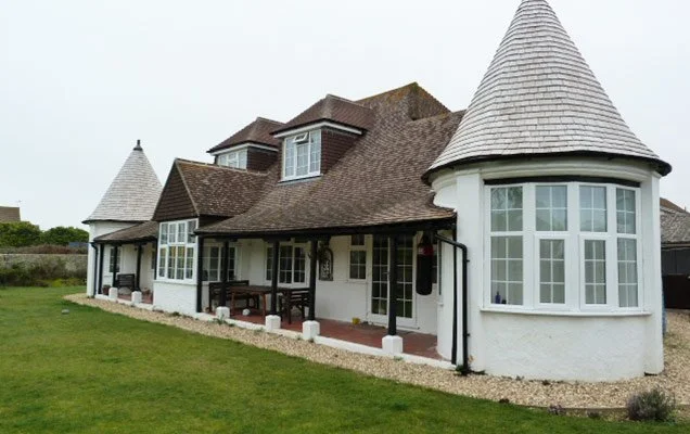 Large house with turret-style towers, white exterior walls, and a brown roof, surrounded by a green lawn and gravel pathway.