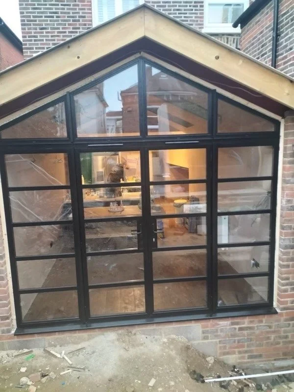 Newly constructed black-framed glass patio enclosure with a gabled roof, attached to a brick building, showing an interior view of the room behind it.