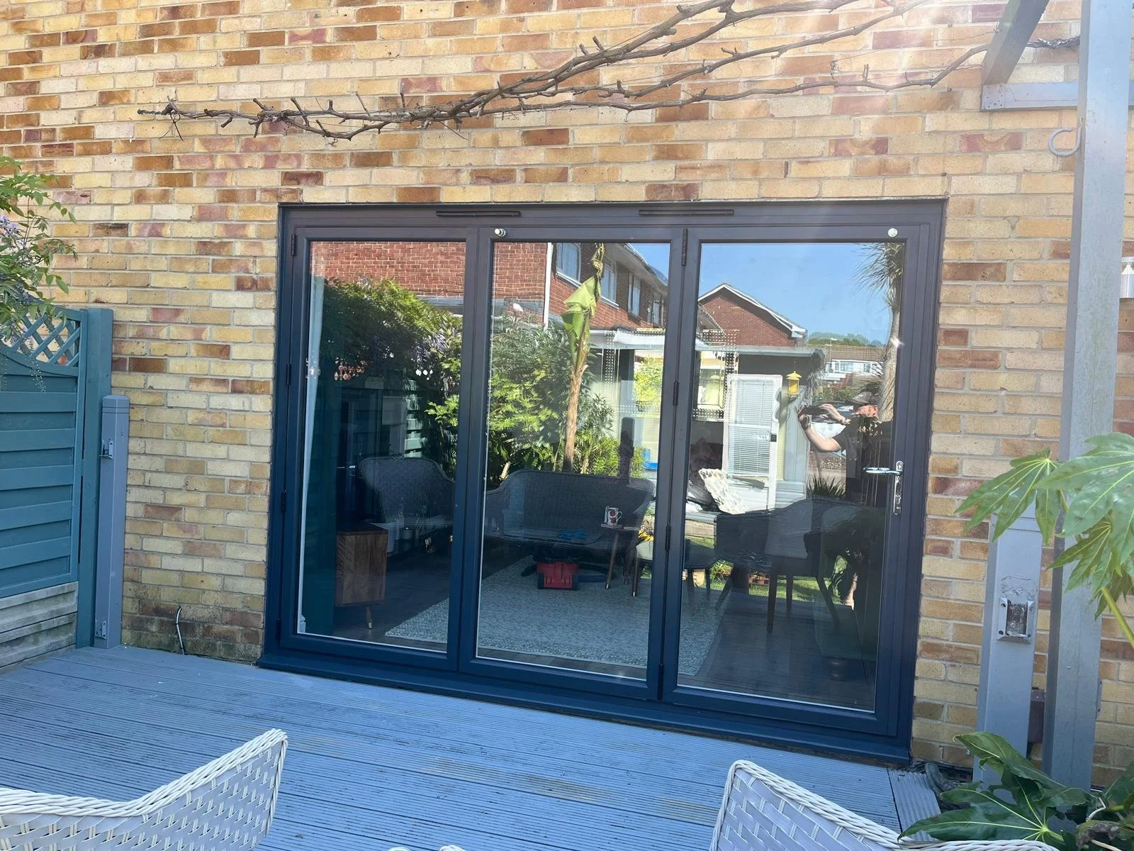 A glass sliding door leading into a patio area with indoor furniture. The patio has a wooden deck with white woven chairs and plants, with a brick exterior wall and a branch above.