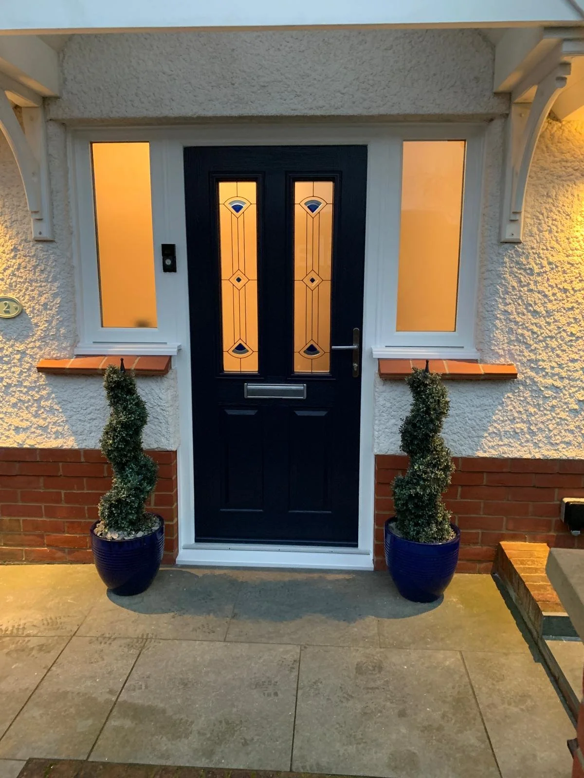 Exterior view of a house front door with decorative glass panels, two potted plants on either side, and warm lighting.