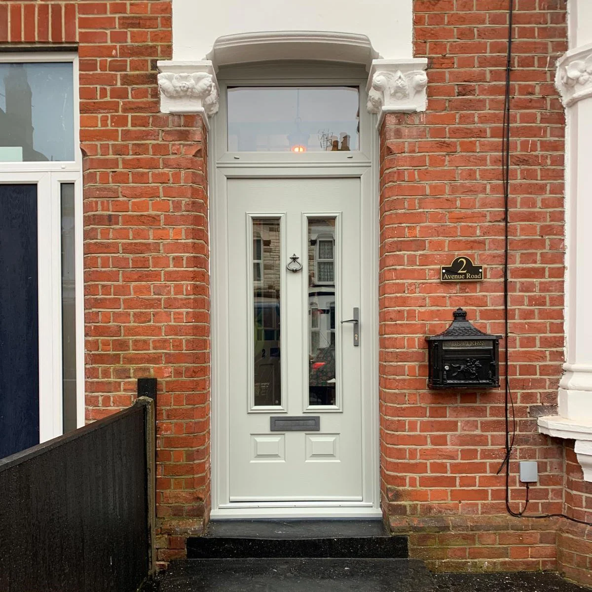 A white front door with glass panels, set in a red brick wall, with a black mailbox to the right and a house number plaque that reads "2 Avenue Road."