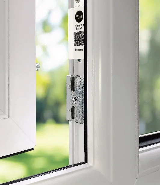 Close-up of a window latch with a Yale smart lock QR code sticker on the frame, outdoors background with trees and sunlight.