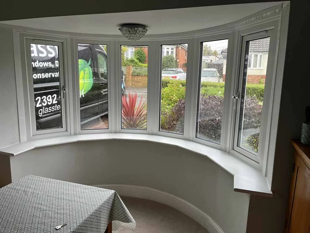 Curved bay window with white frame in a room, overlooking a street with cars and houses outside.