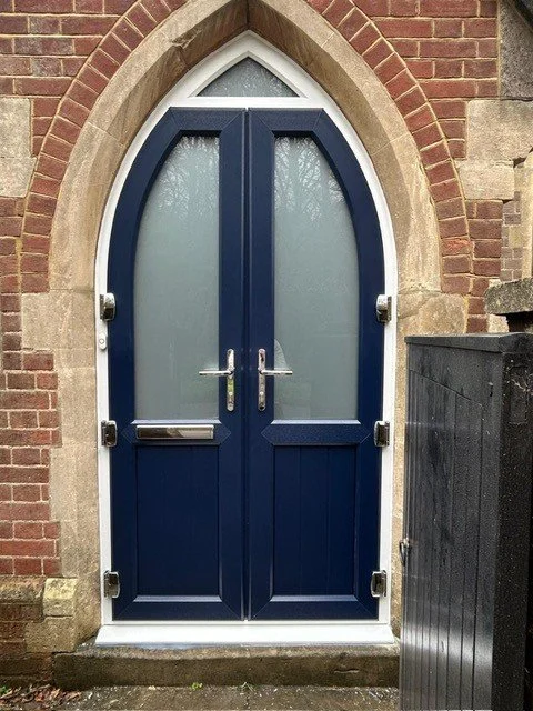 A blue, arched front door with frosted glass panels and silver handles, set in a brick and stone building wall.