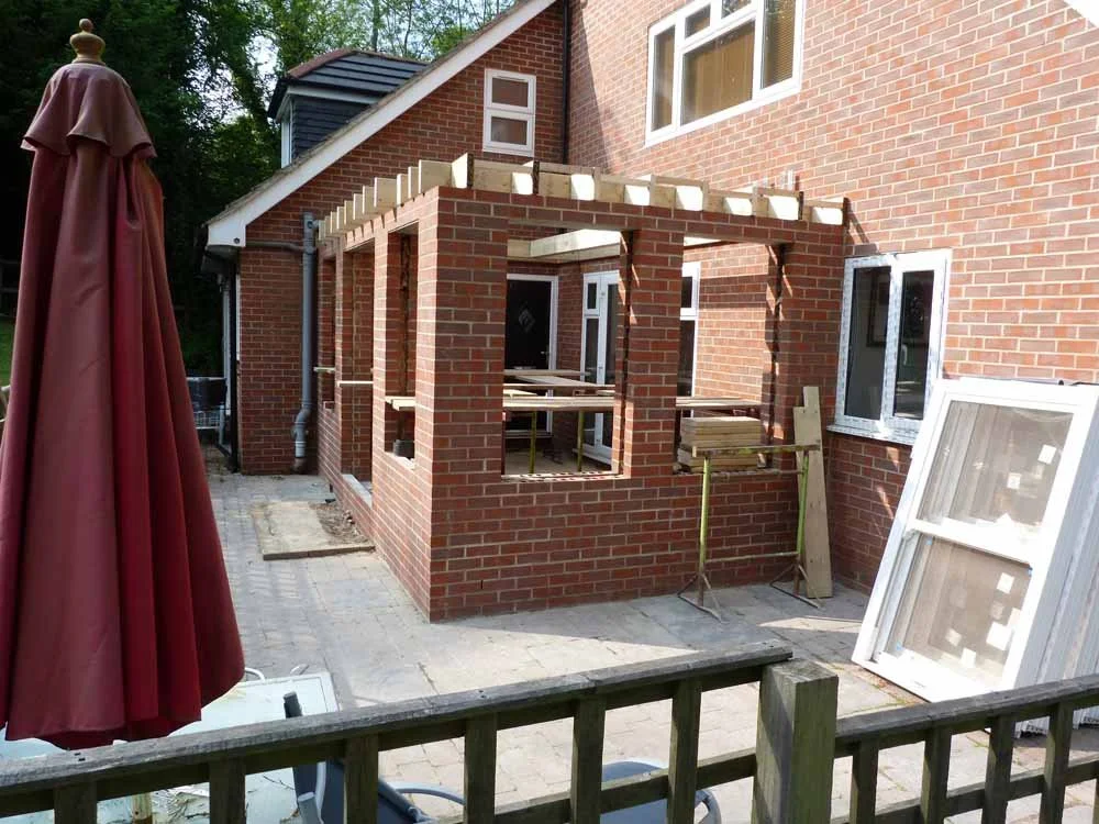 Construction of a brick patio cover or porch on the back of a brick house, with wooden beams and supports in place, and building materials such as window screens leaning against the house.