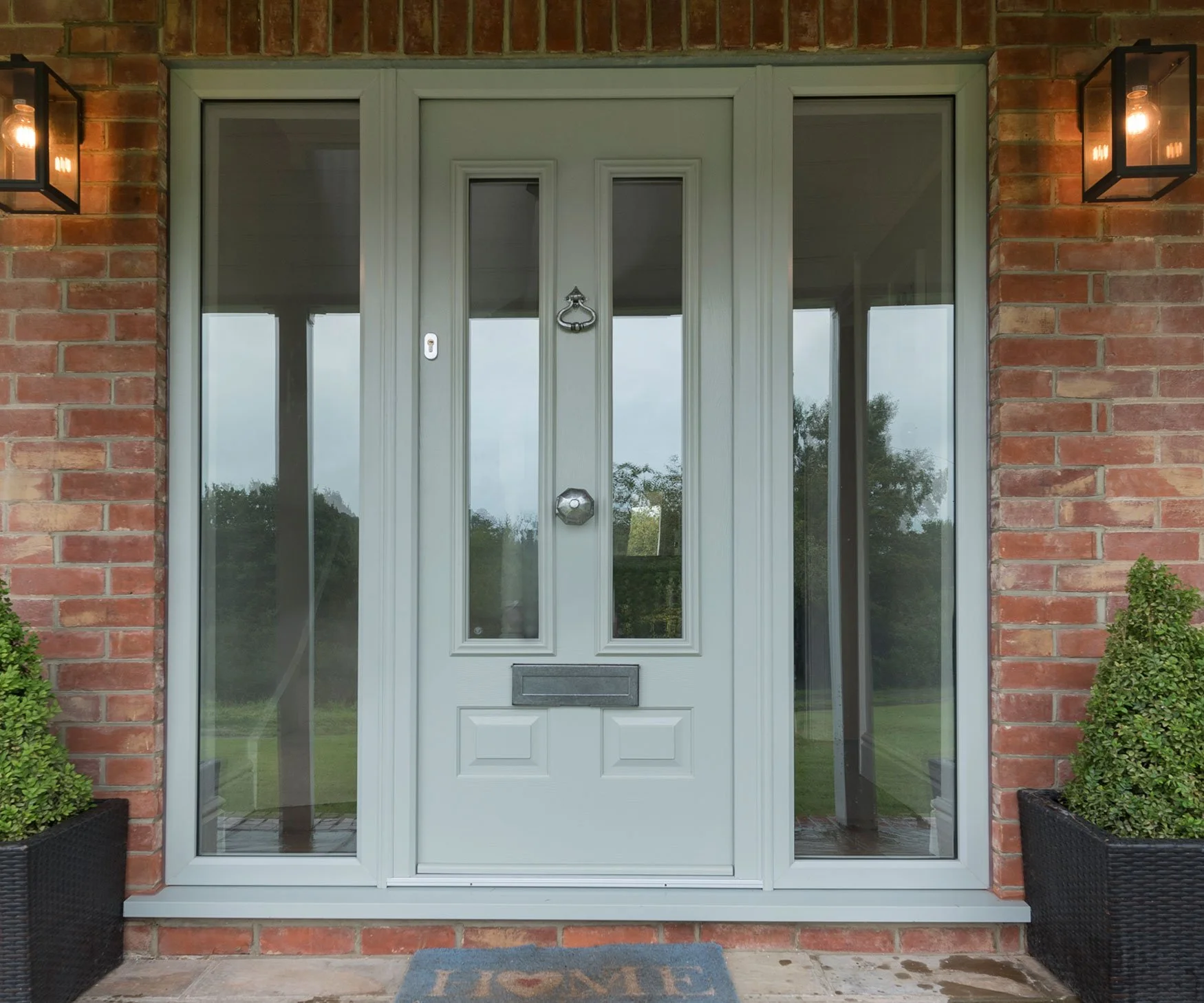 Front door with glass panels on sides, brick wall, two black outdoor wall lanterns, bushes in black planters, and a welcome mat.