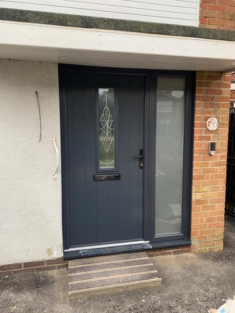 Black front door with decorative glass panel and mail slot, flanked by a large glass window, on a small brick and concrete porch with a wooden step.