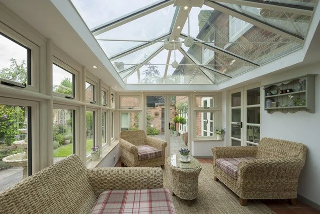 Sunroom with wicker chairs, a glass table, and a glass ceiling, overlooking a garden.