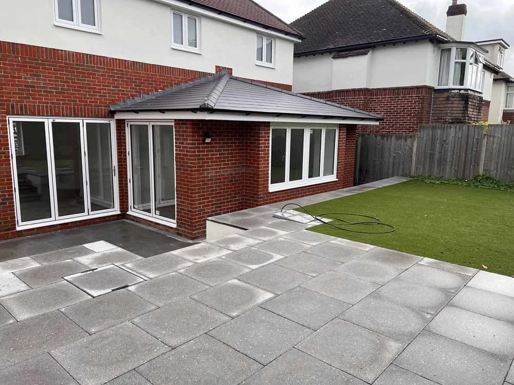 Backyard with patio, artificial grass, red brick house with sliding glass door and windows, fenced yard, and garden hose.