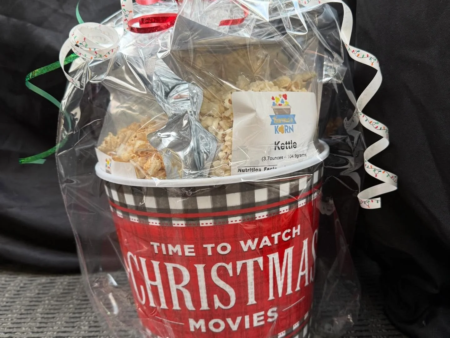 A Christmas-themed popcorn bucket with a red and black checkered design and the words "Time to watch Christmas movies" on it, decorated with curling ribbons, containing popcorn with a bag of caramel popcorn on top.